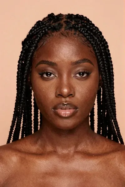 Photorealistic studio portrait of a young Black woman with deep brown skin, full round face, short micro braids with blunt bangs, glossy lips, winged eyeliner, nose rings, soft peach background, direct eye contact, realistic skin texture, high-detail fashion portrait.