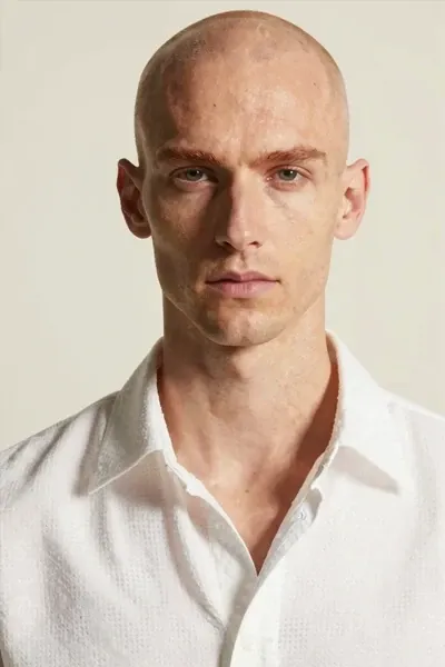 Realistic fashion portrait of a bald man with very fair skin, long narrow face, subtle reddish eyebrows, calm intense gaze, white open-weave shirt, pale cream background, warm directional light, clean minimal composition, detailed skin texture and face contour.