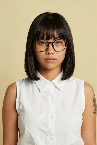 Realistic front-facing portrait of a young Southeast Asian woman with medium tan skin, straight black bob haircut with bangs, large round glasses, calm thoughtful expression, sleeveless white collared top, small earrings, visible arm tattoo, pale yellow studio background, clean editorial lighting.