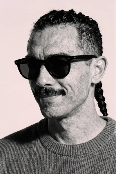 Black-and-white close-up portrait of a lean man with olive skin, sharp cheekbones, thin mustache, undercut hair with one long braid, dark sunglasses, textured knit sweater, confident half-smile, pale pink studio backdrop, strong side light, realistic facial detail.