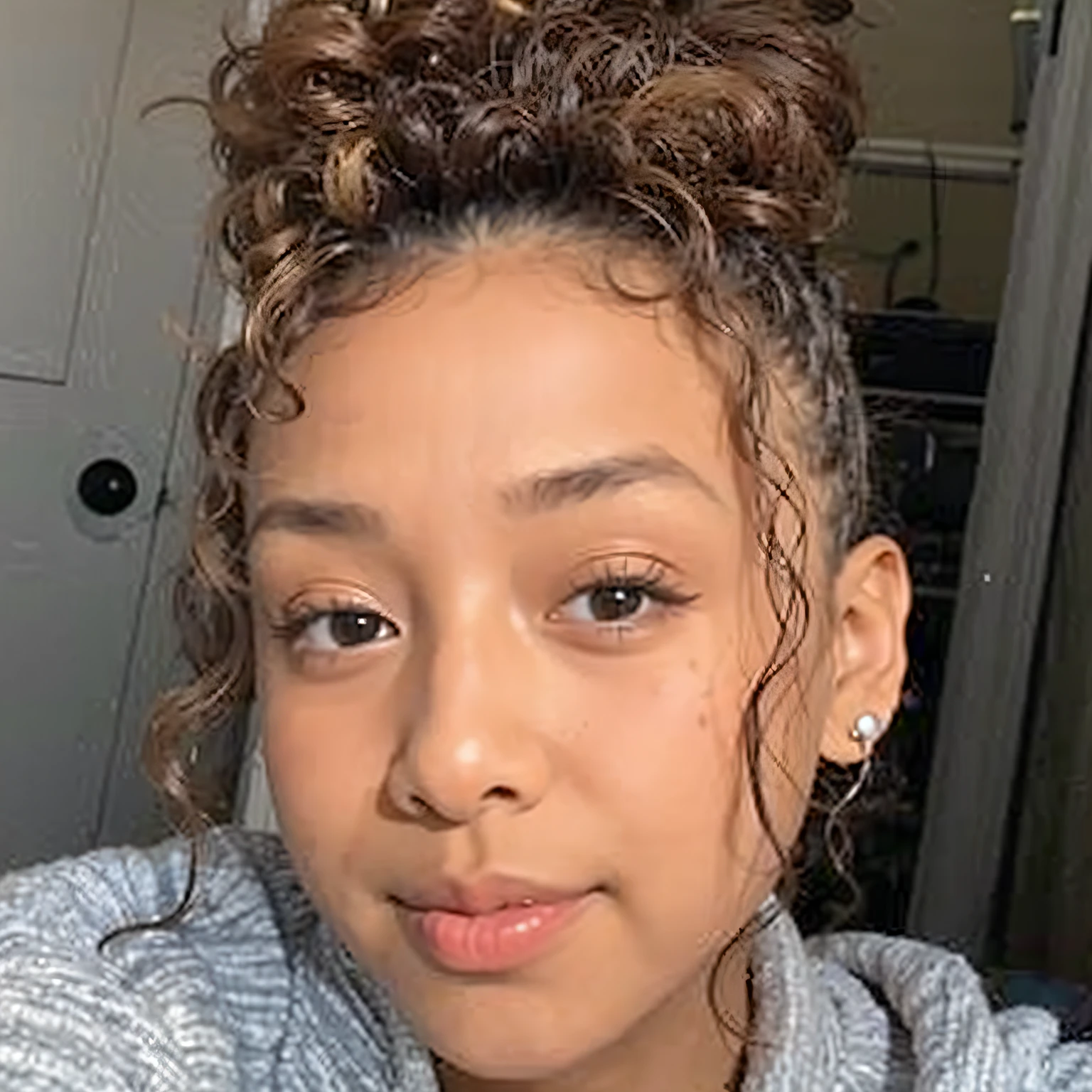 woman with curly hair and a gray shirt taking a selfie, curly messy high bun hairstyle, she is about 1 6 years old, taken in the early 2020s, willow smith young, with dark hair tied up in a bun, light skinned african young girl, she has olive brown skin, her face framed with curls, curly bangs and ponytail