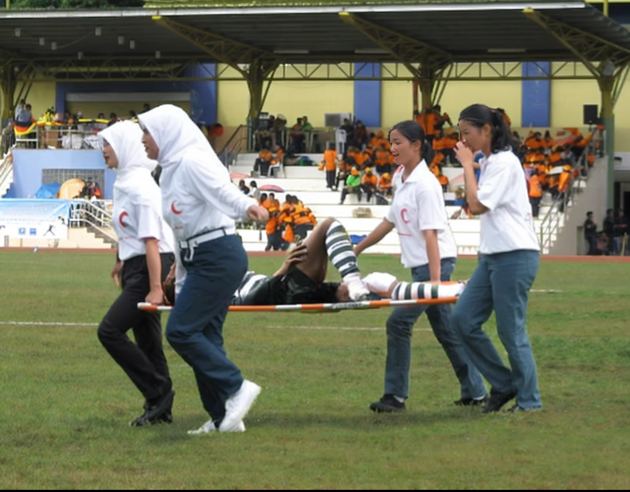 four malaysian girls carry a stretcher, four veiled girls carry a stretcher, a soccer scene in a sports stadium, cool and wet weather conditions, humide ground, rainy sky, injury scene in a sports stadium, stretcher carry, there are four female medics carrying a stretcher, there are four female medics in very shiny coats who are carrying a stretcher in a sports stadium, there is a wounded male soccer player in a matte short cotton sports outfit lying on the stretcher, an injured male soccer player in matte cotton sportswear is lying in pain on a stretcher, a soccer player in matte cotton sports clothes is rearing up in intense pain while lying on a stretcher, dramatic scene, theatralic posing scene, dramatic pity scene, injury soccer, first aid, help, pity, there are four female medics in wetlook high-shine coats who are looking very sad and very terrified and very shocked, the injured soccer player is screaming out in pain while he is carried from the pitch on a stretcher 