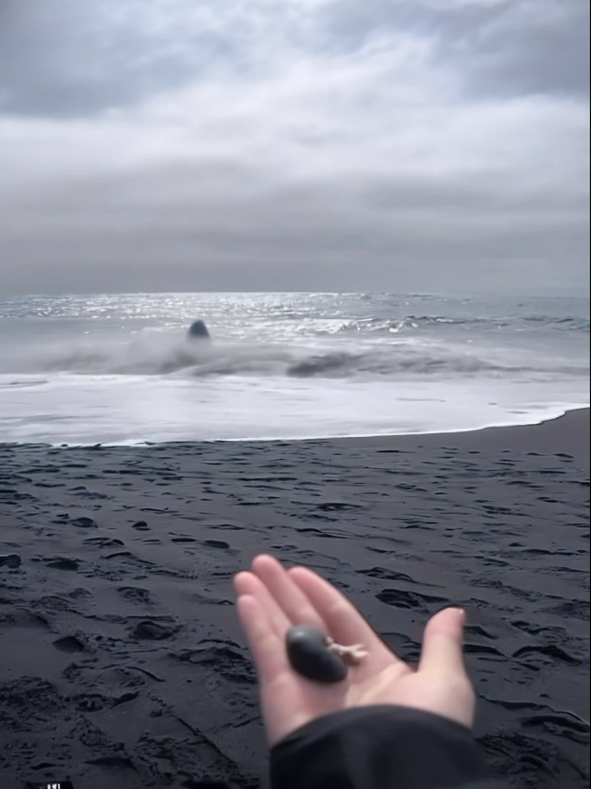 Someone on the beach with a small thing in their hands, Black sand, wave a hand at the camera, levitating sand, the ocean in the background, the ocean in the background, maybe small waves, video still, Strange suspension stone, The sea in the background, realistic ocean, Ferrofluids ocean, In the calm waters of Iceland, middle of the ocean