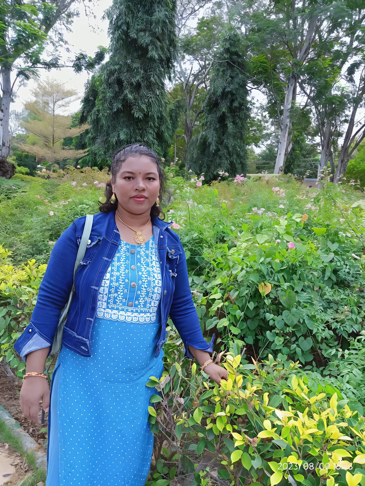 woman in blue dress standing in a garden with bushes and trees, standing in a botanical garden, 30-year-old woman from cuba, taken in the early 2020s, photo taken in 2 0 2 0, in garden, she is the center of the garden, ancient garden behind her, amidst nature, in red gardens, background is heavenly