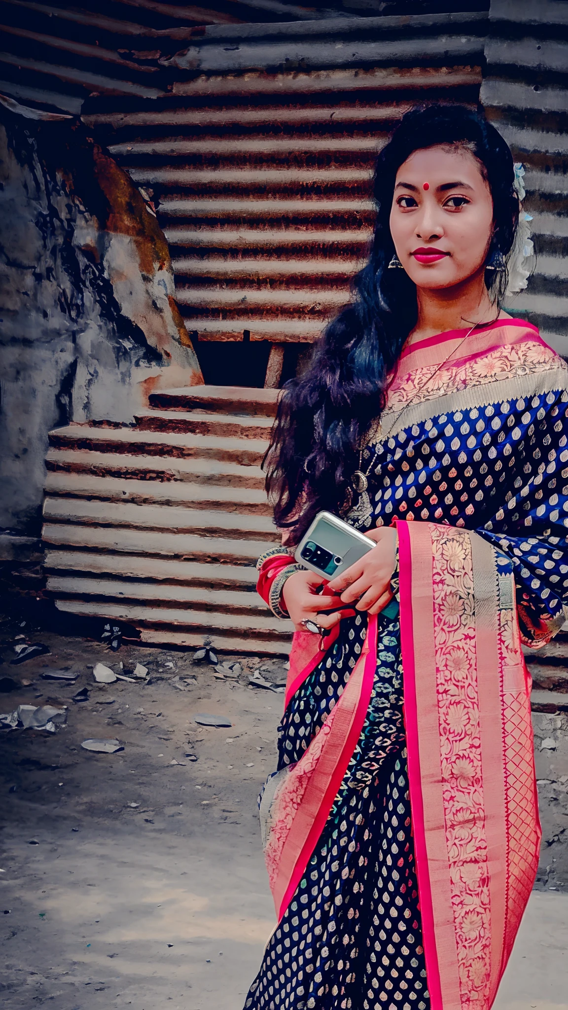 woman in traditional sari holding a cell phone in front of a corrugated wall, traditional beauty, wearing a sari, dressed in a sari, with lovely look, wearing sari, blue colored traditional wear, candid picture, assamese aesthetic, wearing bihu dress mekhela sador, shot on nikon z9, traditional clothes, candid photo, looking from side