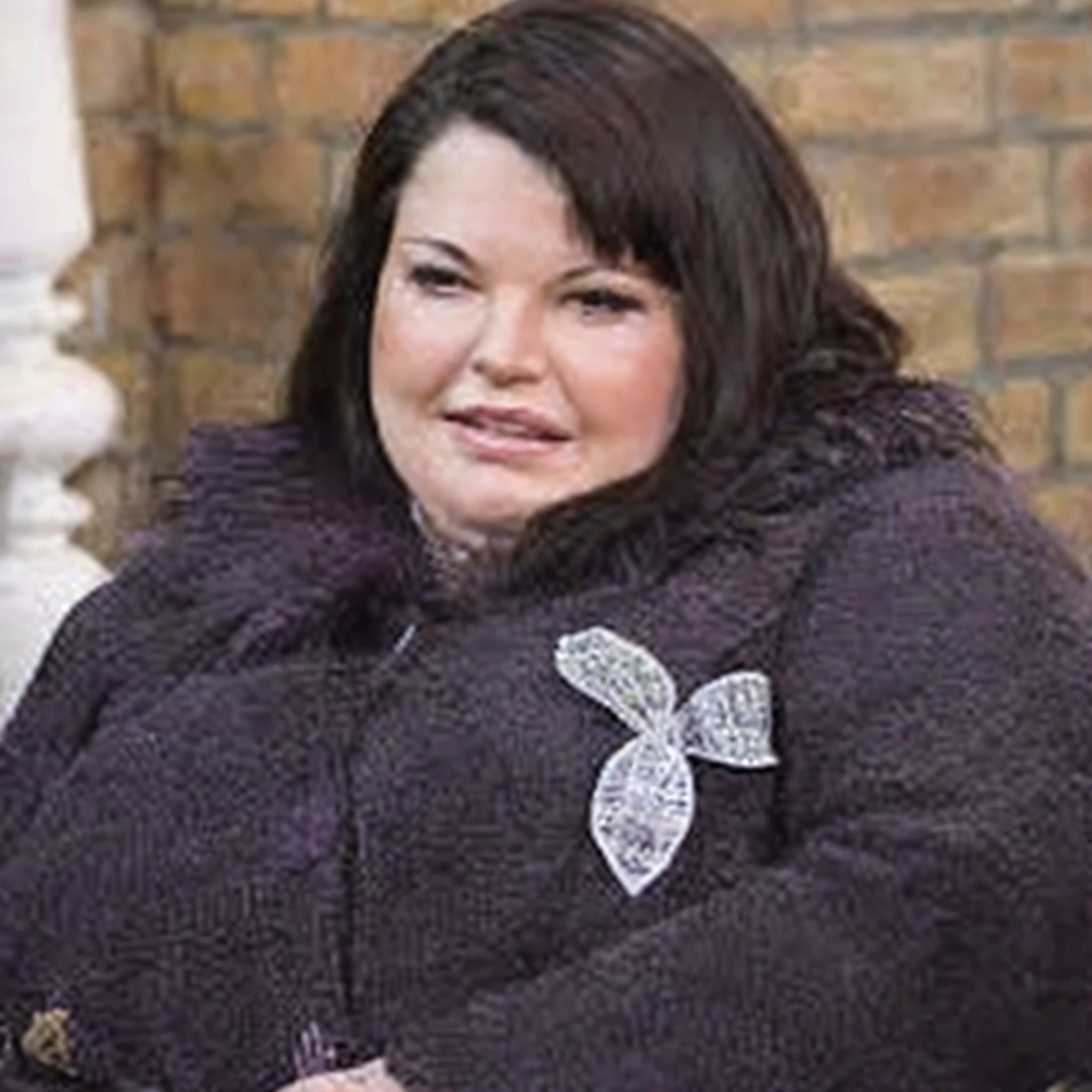 Woman in purple coat sitting on a bench with a brick wall behind her, taken in the late 2010s, taken in the early 2020s, inspirado em Lorraine Fox, mulher gorda, mulher feia, Vivian Westwood, Um retrato de uma mulher rechonchuda, her black hair is a long curly, Kimberly Asstyn