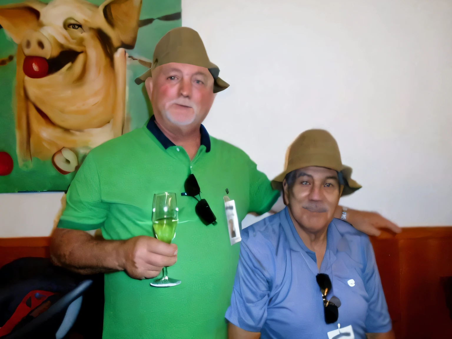 On the wall is the head of a pig, In front are two men in hats, one is seated, The one standing has a glass of champagne in his hand