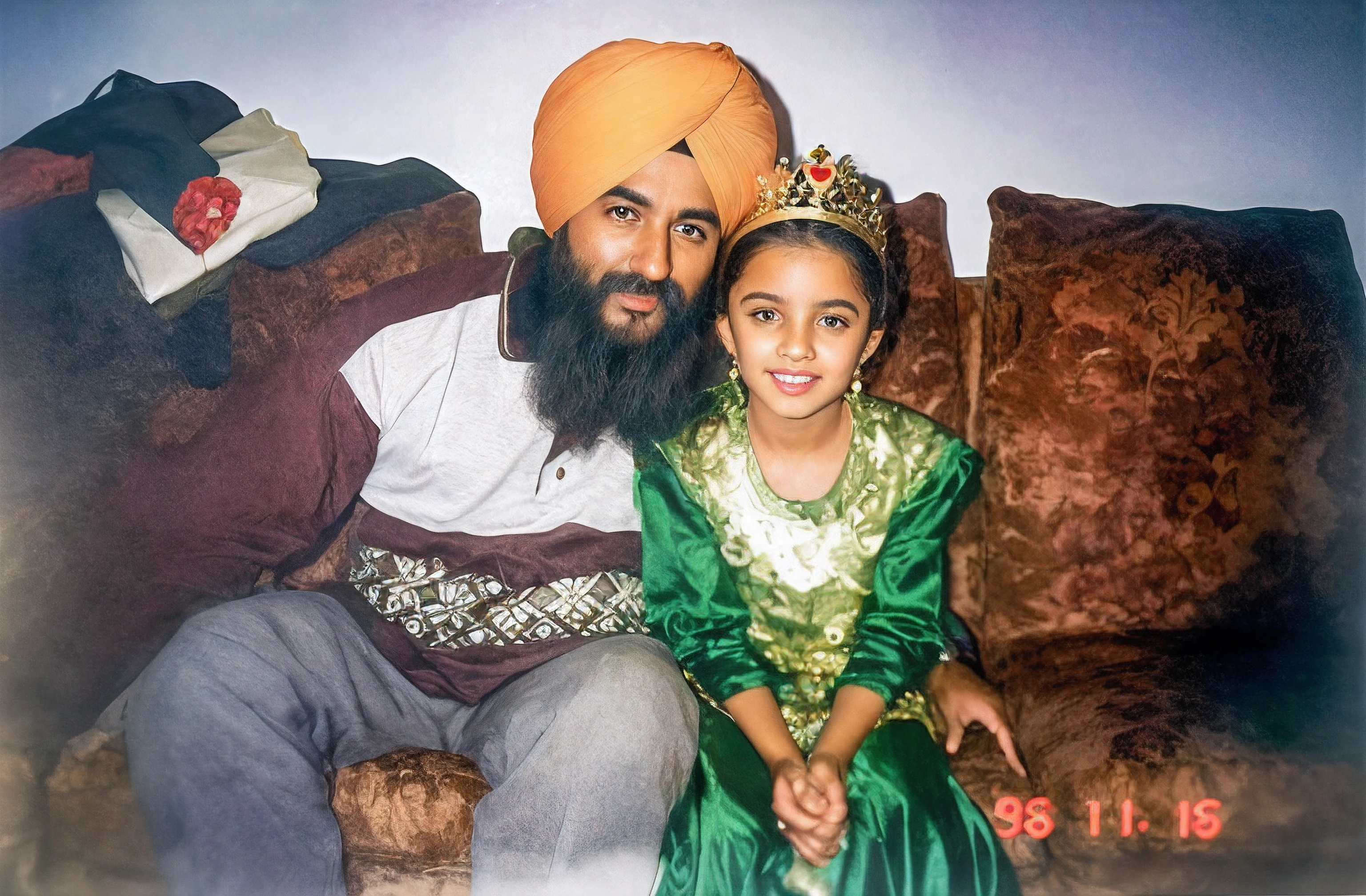 There's a man and a girl sitting on a couch, homem com barba grande de cor preta, both Muslims, turbante amrelo, sofa castanho com almofadas, Girl with golden crown with a red heart in the center, vestido verde, raw photo, amazing photograph, best quality, 8K, high quality, Ultra detailed, (best quality), (high detailed skin, full sharp:1.2), 8k uhd, dslr, detailed face