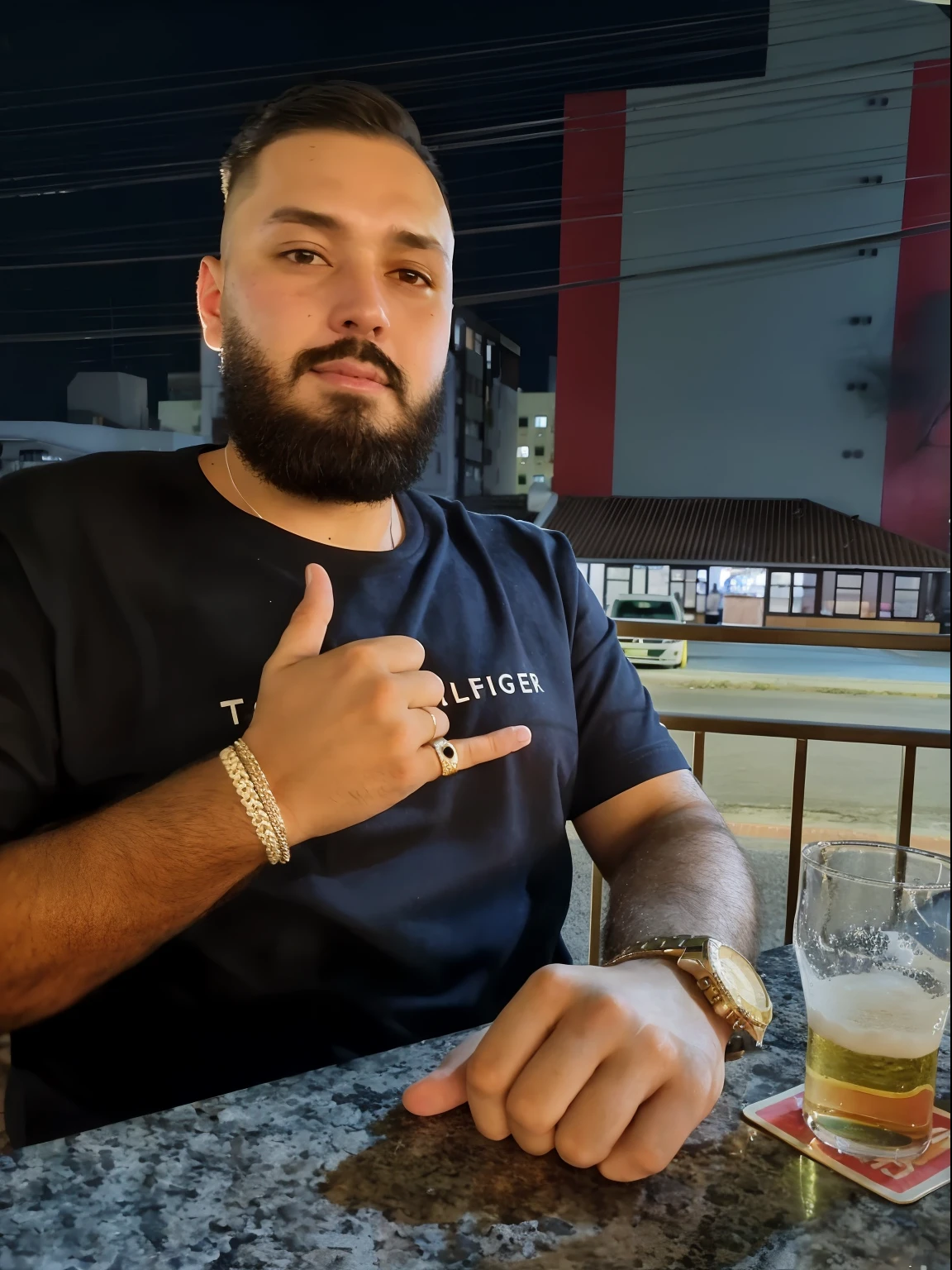 There's a man sitting at a table with a glass of beer, em segundo plano imagem borrada com predios atraz dele.