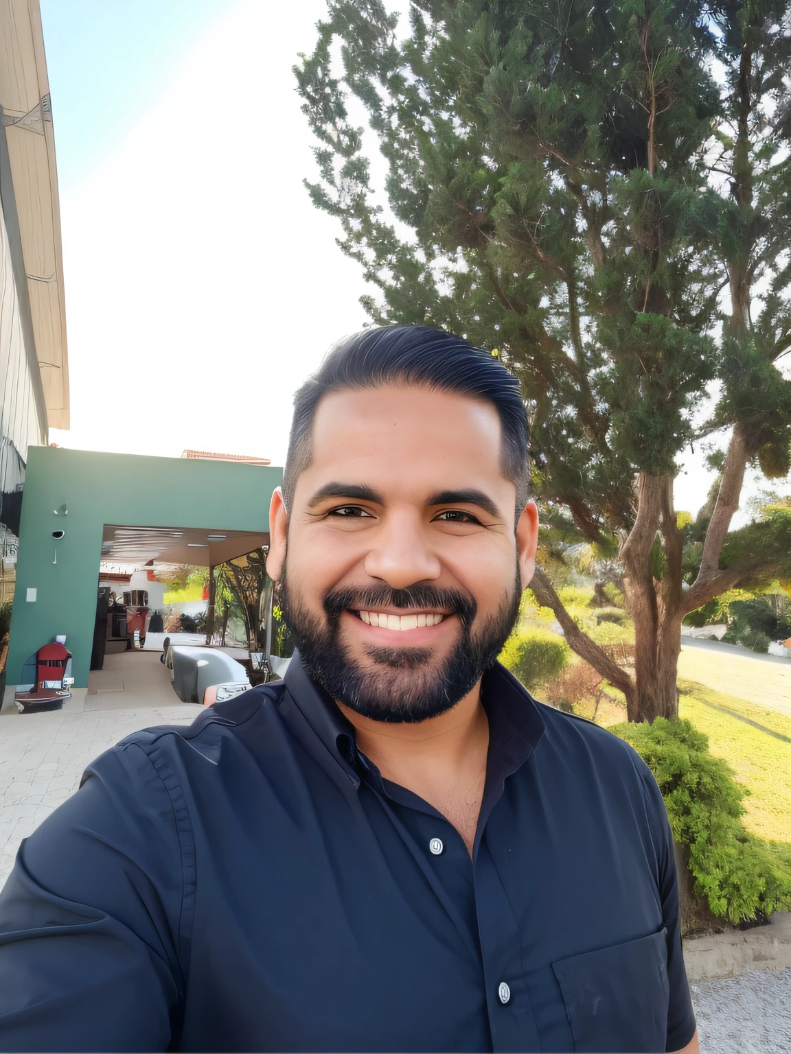 A man stands outside a building with a tree in the background, jardim, He's dressed in a black shirt, sorrindo, com barba preta. Tem o cabelo preto penteado para o lado. He's got black eyes and he's taking a selfie.