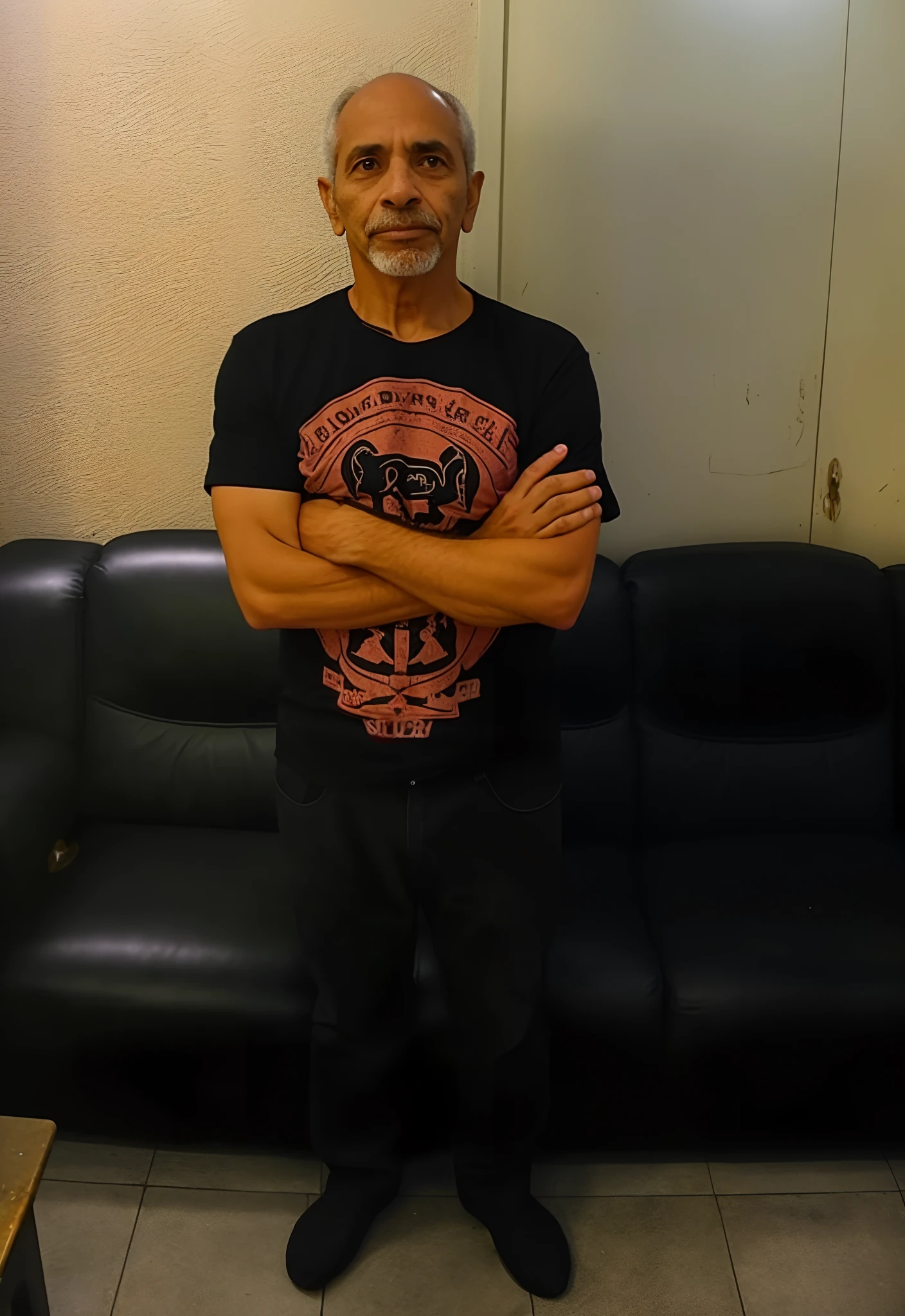Arafed man standing in front of a black sofa with arms crossed, he is wearing a black t-shirt, ele tem cerca de 6 0 anos de idade, ele tem cerca de 60 anos, ele tem cerca de 7 0 anos de idade, ele tem cerca de 5 0 anos de idade, ele tem cerca de 50 anos, standing in a dimly lit room