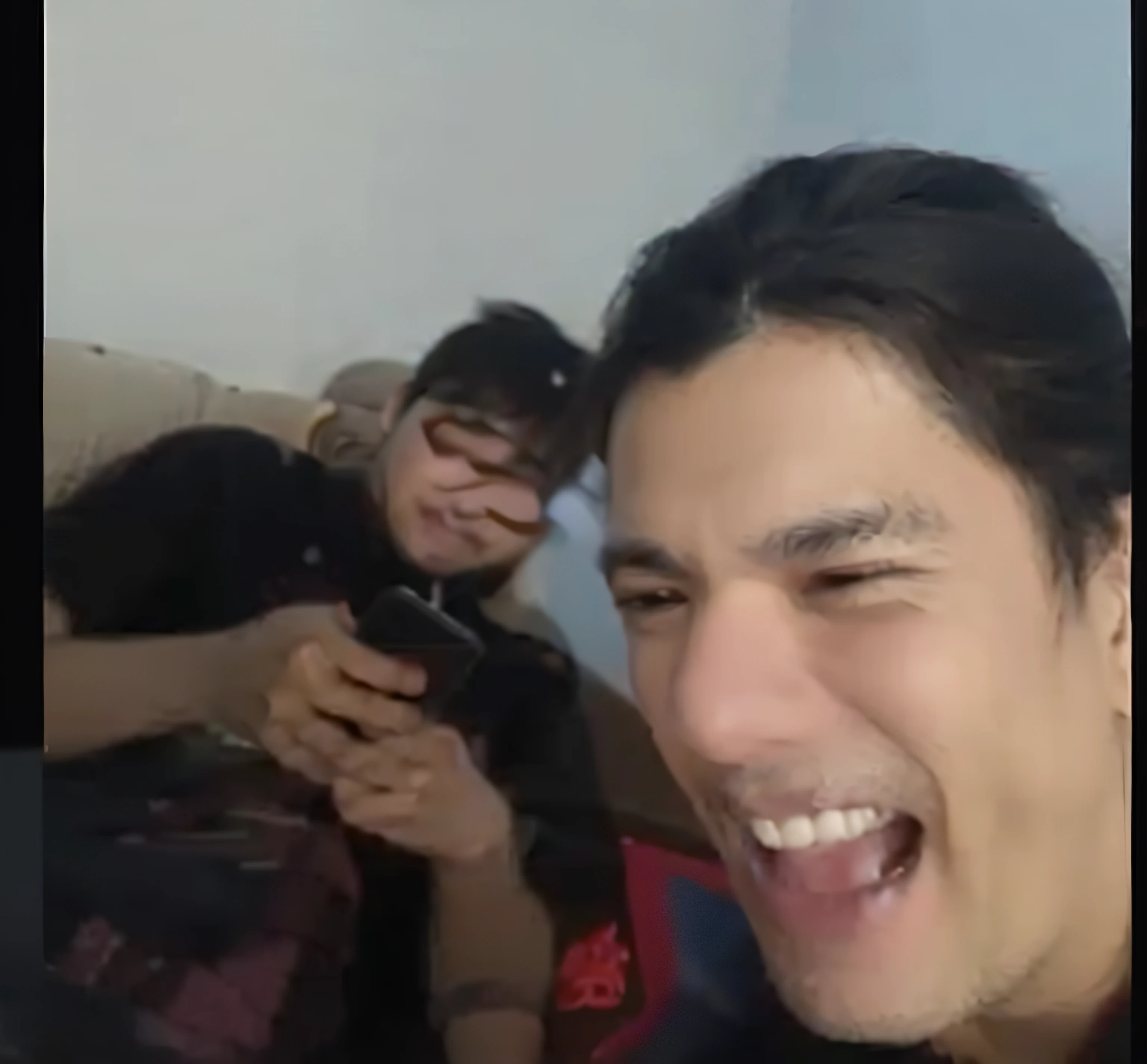 One person in the back fiddling with his phone and sitting on the couch while the other is in front with his phone in his hand and recording video laughing at him