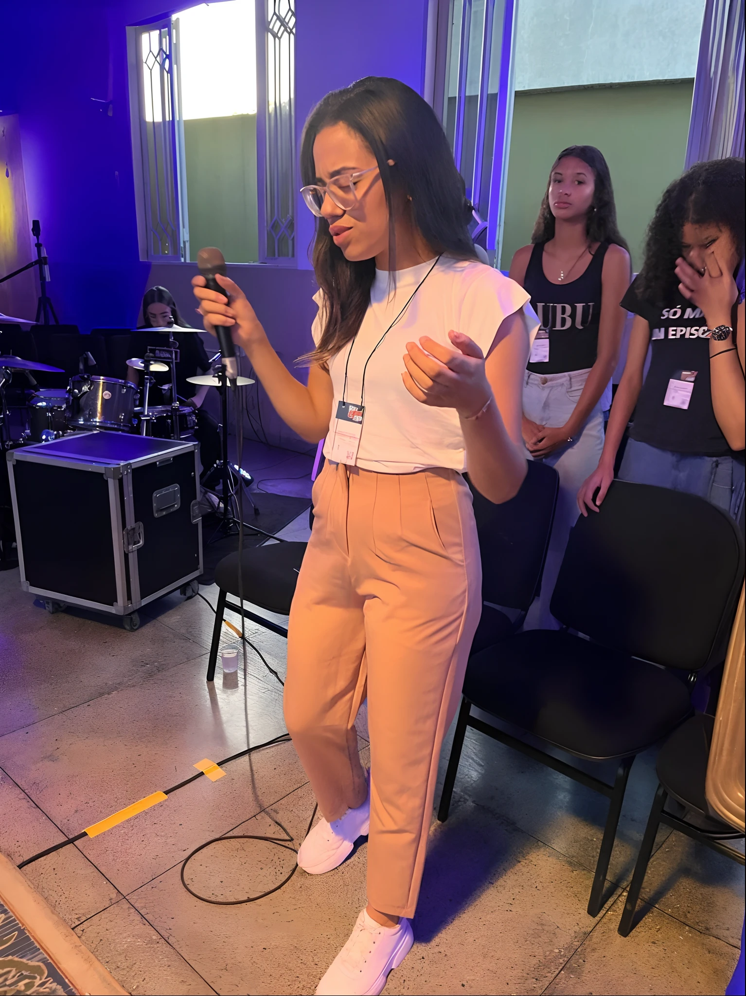 There's a woman standing in front of a microphone and a microphone, Apresentando-se no palco, Executando um videoclipe, no palco, Executar, olhar infantil, em uma regata branca cantando, taken in the early 2020s, classic vibes, vibing to music, suave em _ O pano de fundo, nostalgic vibes, dancing in the background, segurando um microfone