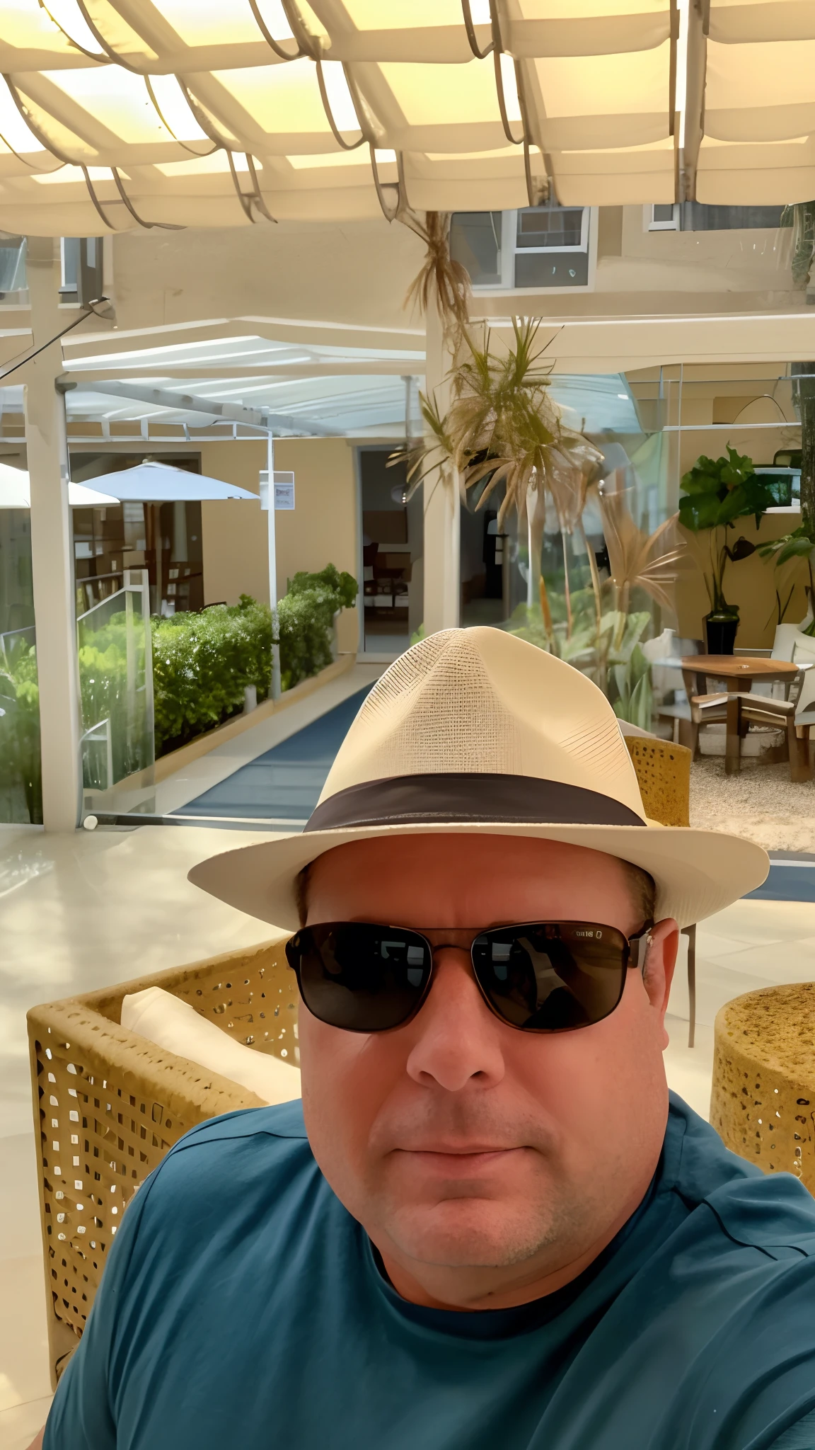 There is a man wearing a hat and sunglasses sitting at a table, wearing sunglasses and a hat, wearing a travel hat, Gorjetando seu fedora, sentado em um lounge, he is wearing a hat, usando um parker, ao lado de uma piscina tropical, wearing sunglasses and a cap, ao lado de uma piscina, com palmeiras nas costas, on the terrace