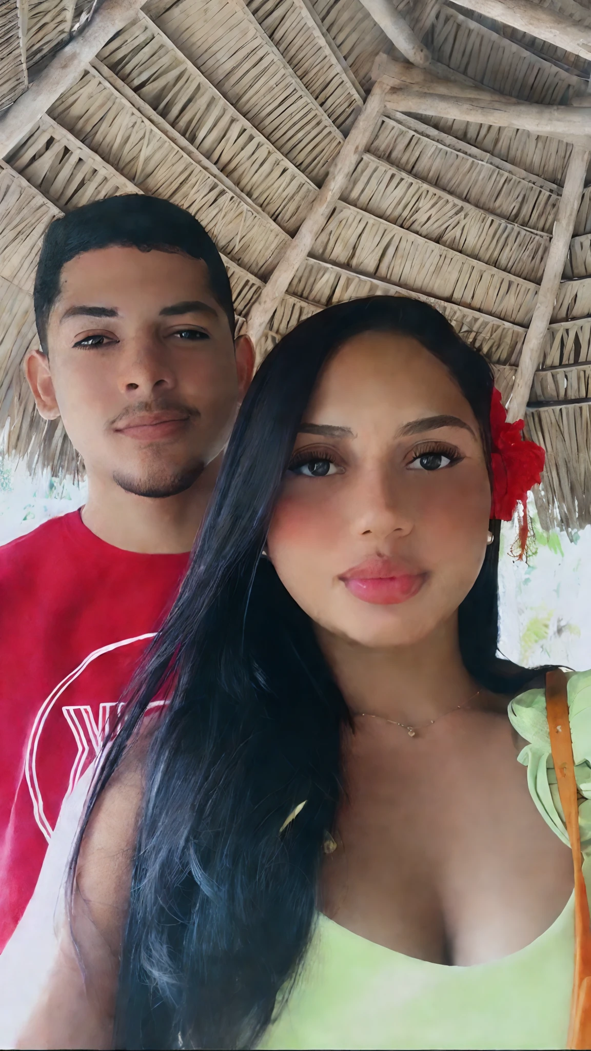 There's a man and a woman under a thatched roof, Mulher de 23 anos cabelos escuro flor vermelha na orelha e blusa verde, pele parda. Homem branco com camisa vermelha, cabelo preto bochechas rosadas