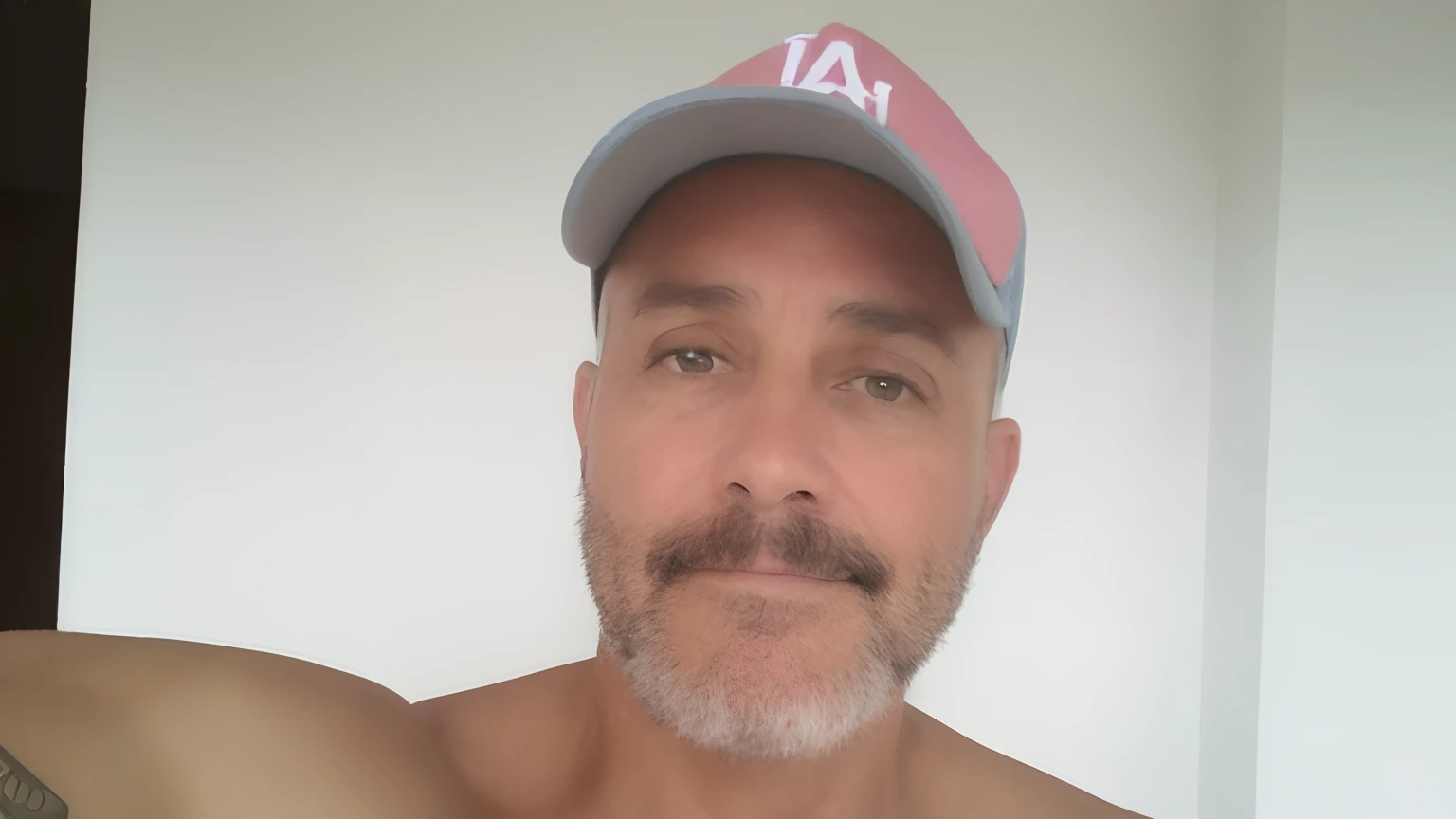 Homem de 49 anos, cabelo e barba curtos e grisalhos. Wears a baseball-style cap.