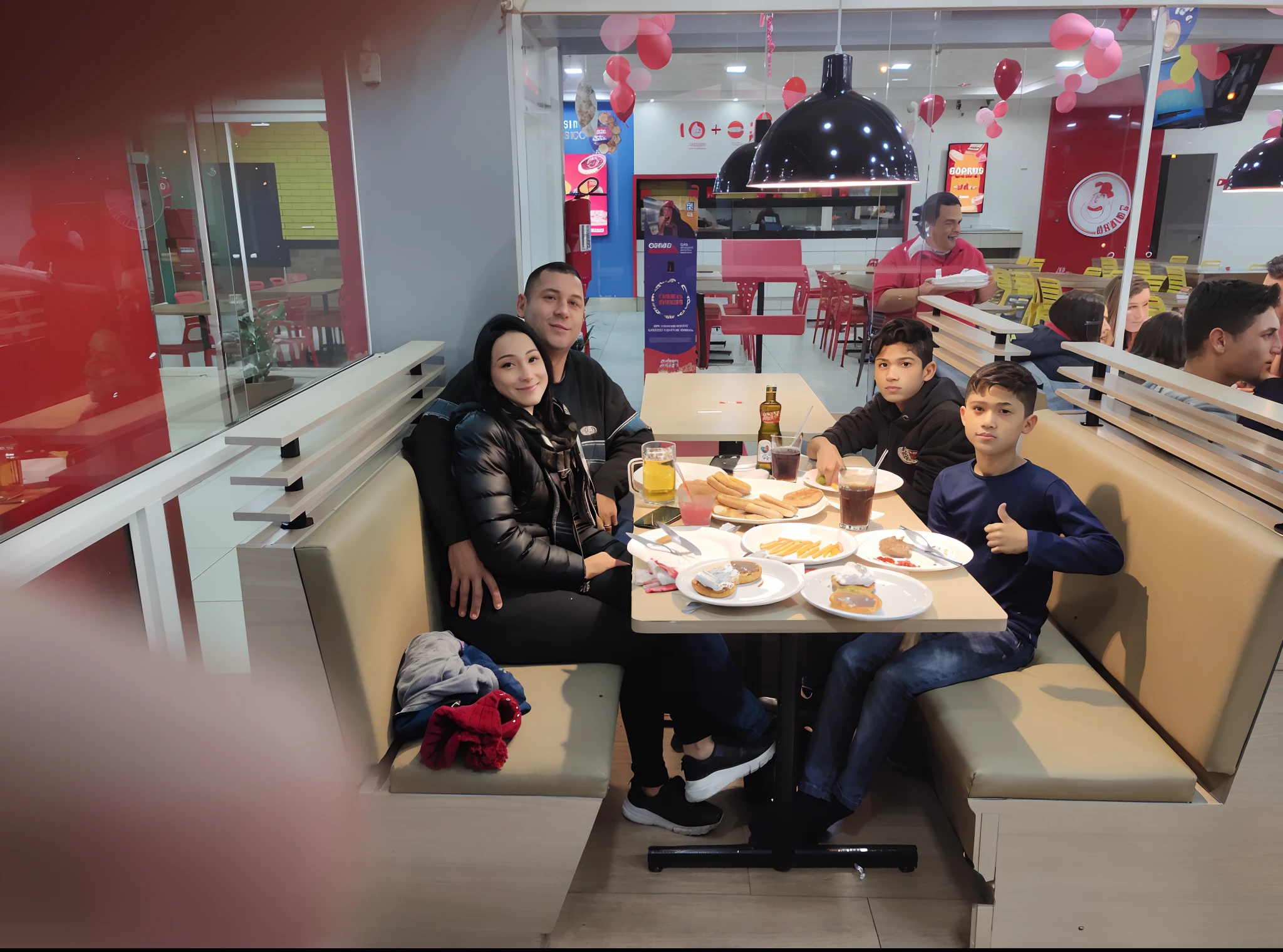 Woman in black jacket hugging man in black coat with a blue stripe in the middle of the coat,  A boy making OK sign in jeans next to another boy in black coat, Everyone is seated with the table with 4 plates with open sfirras