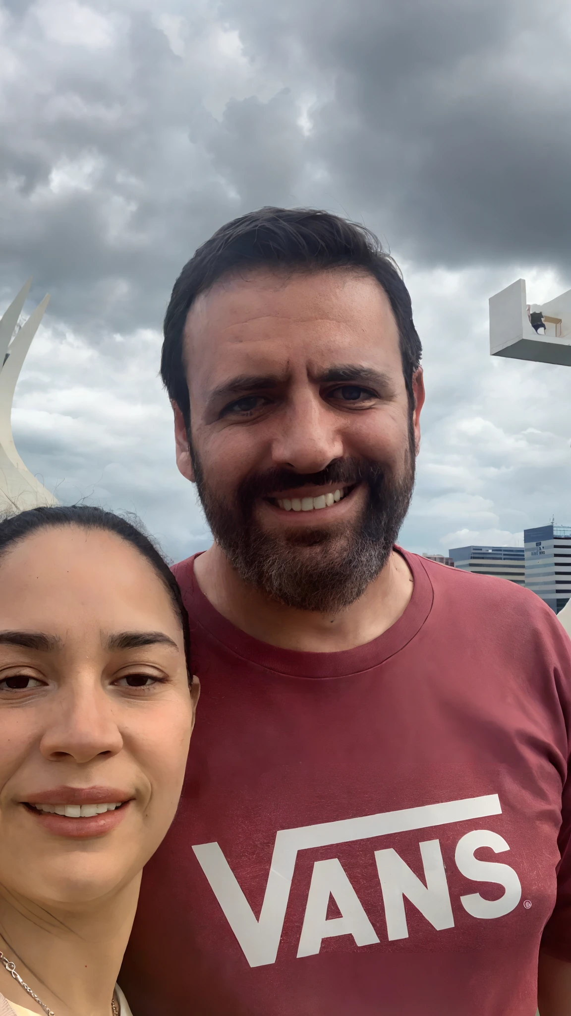 There is a man and a woman together in front of a building, Fotografia selfie 8k, In Sao Paulo, imagem de perfil, vacation photo, imagem de qualidade muito baixa, com montanhas ao fundo, foto de perfil headshot, imagem do avatar, bom tempo nublado leve, Karla Ortiz, com uma cidade em segundo plano, foto do perfil, standing on a skyscraper rooftop