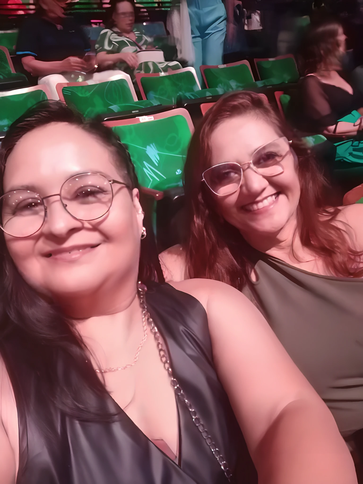 There are two women sitting Pixar style in a stadium with green seats, audience selfie, both smiling for the camera, em Las Vegas, audience in the background, blurry, imagem de qualidade alta, foto do concerto, teatral!, imagem de perfil, Em Matinee estreia, non blurry, vestindo verde, mulheres bonitas, pessoas curtindo o show, belos arredores