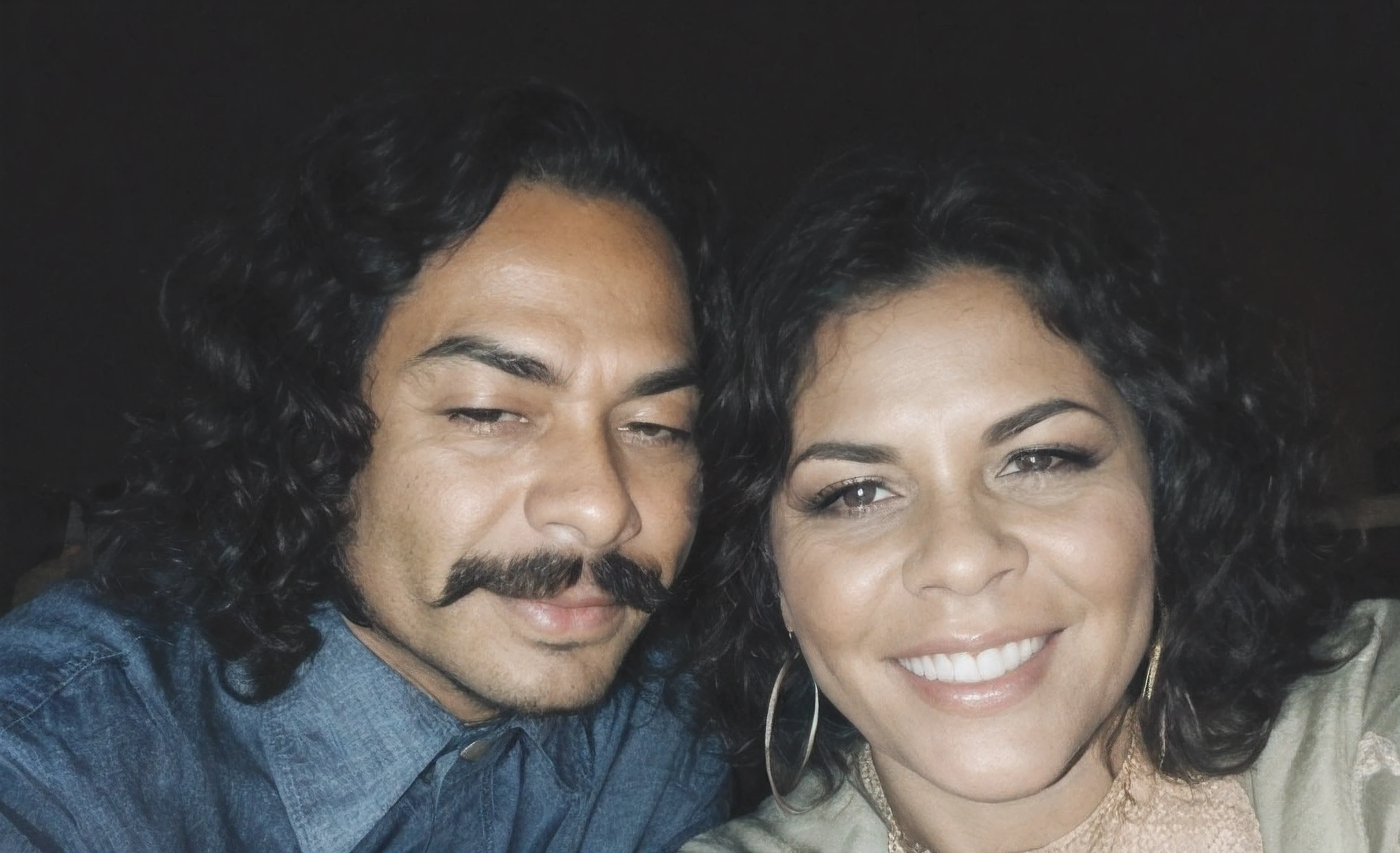There is a man and a woman sitting at the table with a beer, momma and papa, homem de cabelo preto encaracolado comprido com bigode preto, camisa azul de manga curta, pele escura, mulher de cabelo preto encaracolado, pele negra, sorriso, olhos castanhos, com um vestido de tecido verde, branco e roxo de manga curta