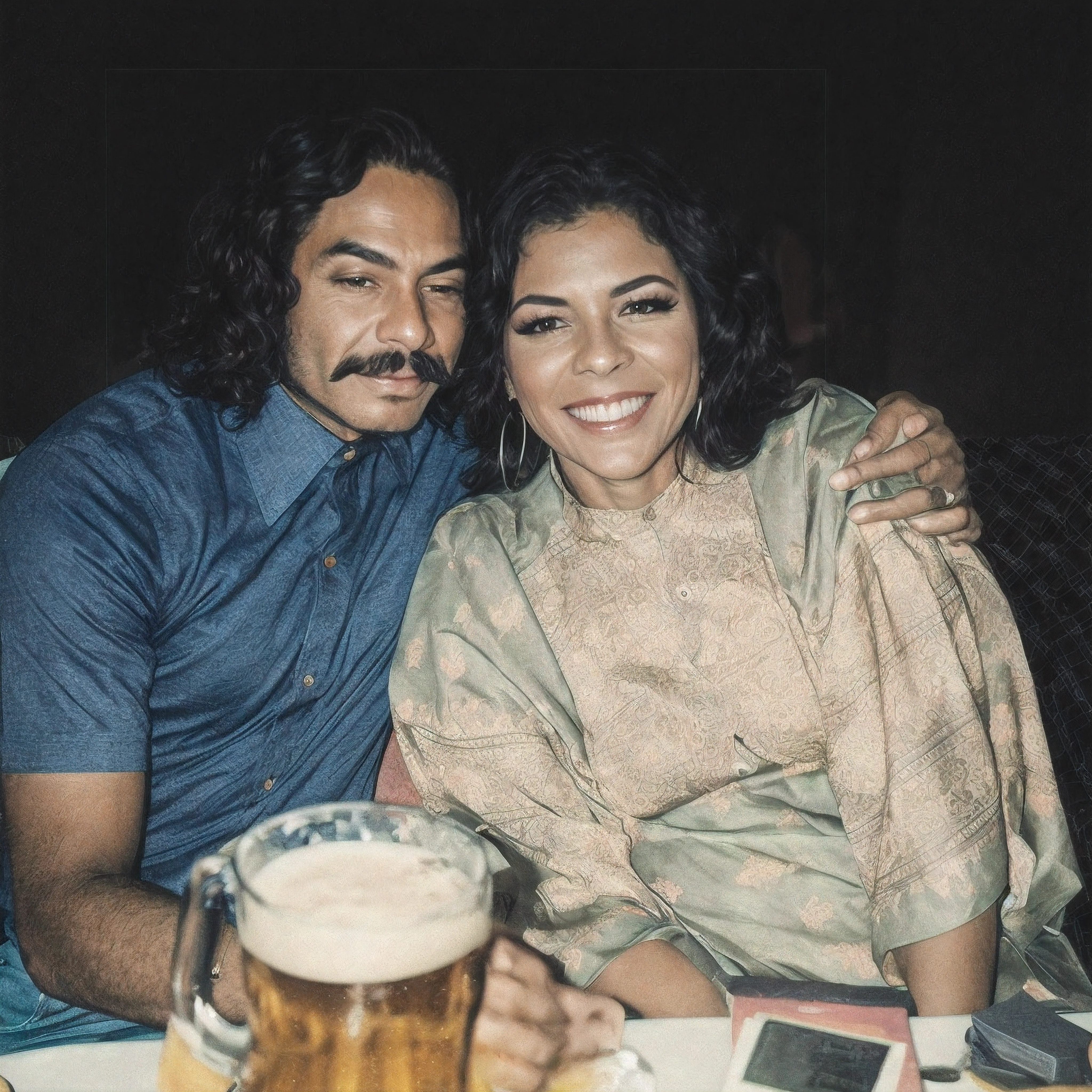 There is a man and a woman sitting at the table with a beer, momma and papa, homem de cabelo preto encaracolado comprido com bigode preto, camisa azul de manga curta, pele escura, mulher de cabelo preto encaracolado, pele negra, sorriso, olhos castanhos, com um vestido de tecido verde, branco e roxo de manga curta, raw photo, amazing photograph, best quality, 8K, high quality, Ultra detailed, (best quality), (high detailed skin, full sharp:1.2), 8k uhd, dslr, detailed face