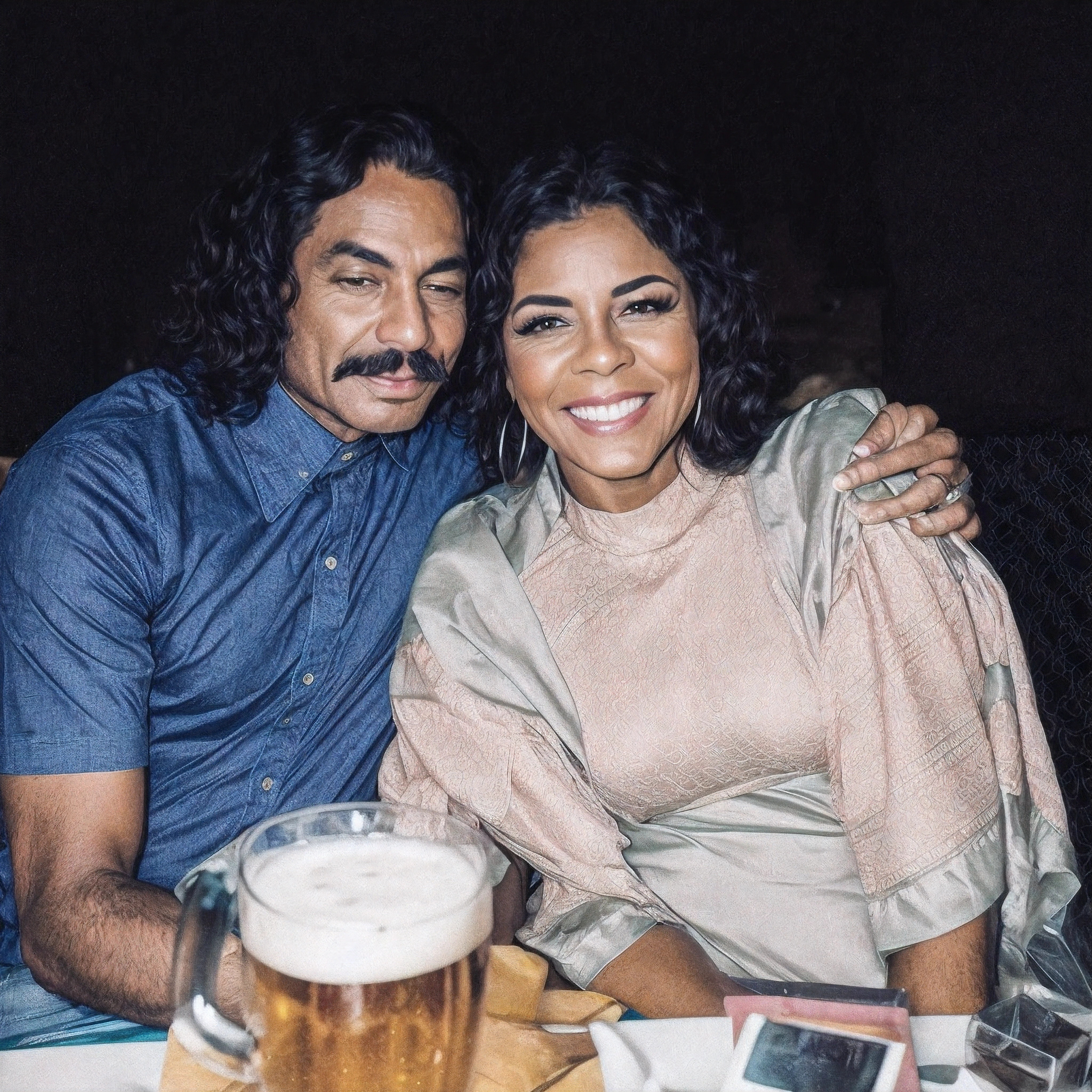 There is a man and a woman sitting at the table with a beer, momma and papa, homem de cabelo preto encaracolado comprido com bigode preto, camisa azul de manga curta, pele escura, mulher de cabelo preto encaracolado, pele negra, sorriso, olhos castanhos, com um vestido de tecido verde, branco e roxo de manga curta, raw photo, amazing photograph, best quality, 8K, high quality, Ultra detailed, (best quality), (high detailed skin, full sharp:1.2), 8k uhd, dslr, detailed face
