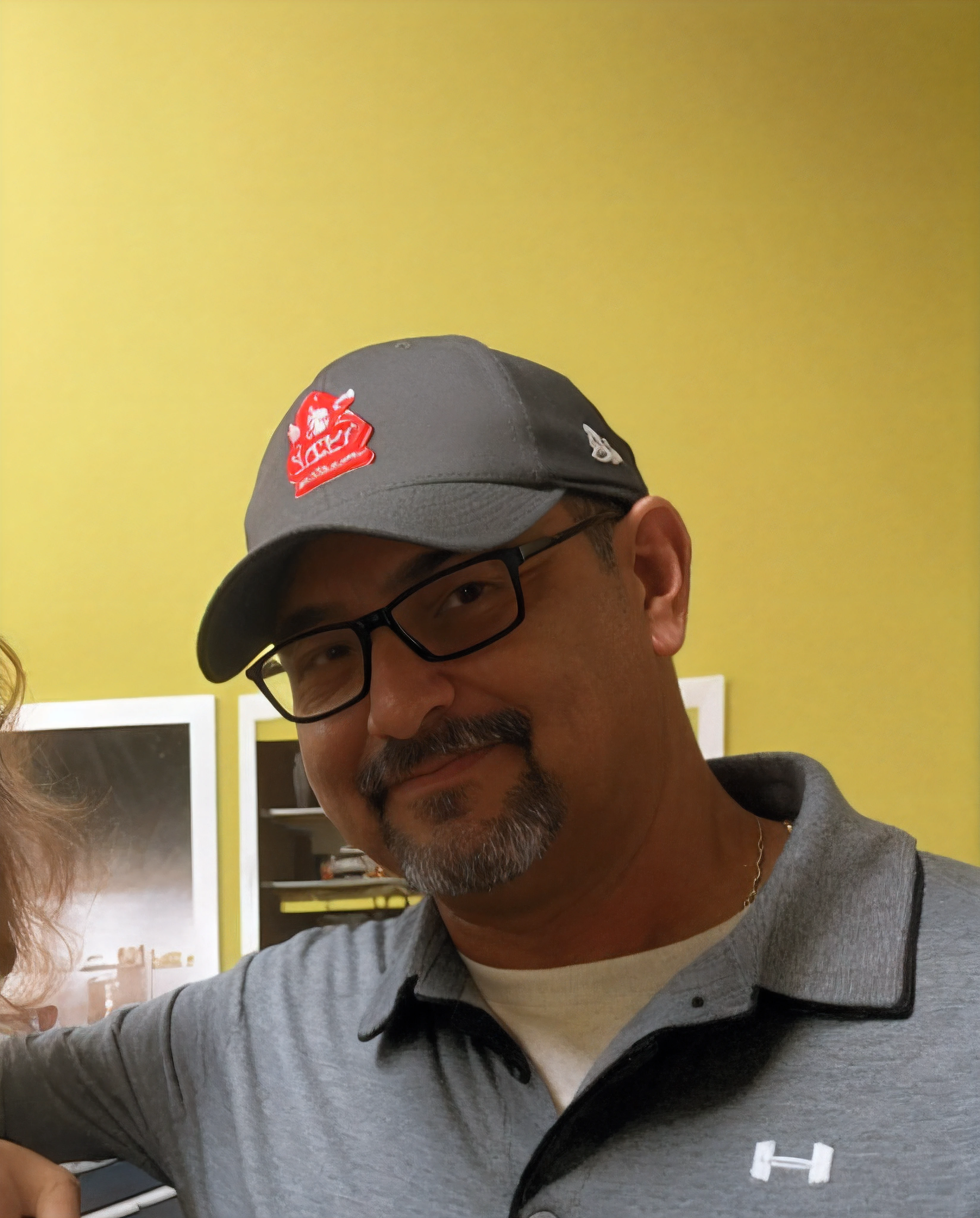 Homem de 45 anos, black hat with a red letter, Black Rectangular Glasses, olhos negros, barba preta grisalha, has a white sweater and a gray polo shirt with a white symbol