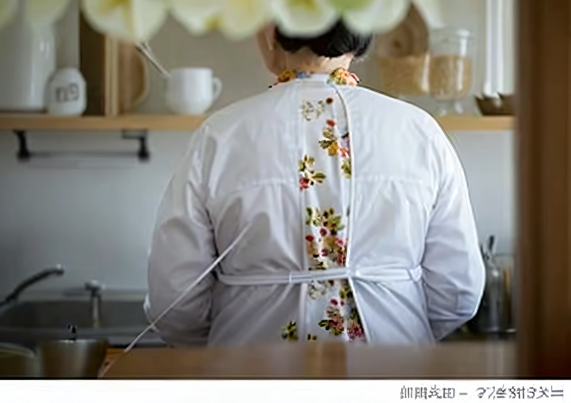 A woman in her 80s standing in the kitchen, She is wearing a white coat and a bright floral blouse。., her back is turning, back - shot