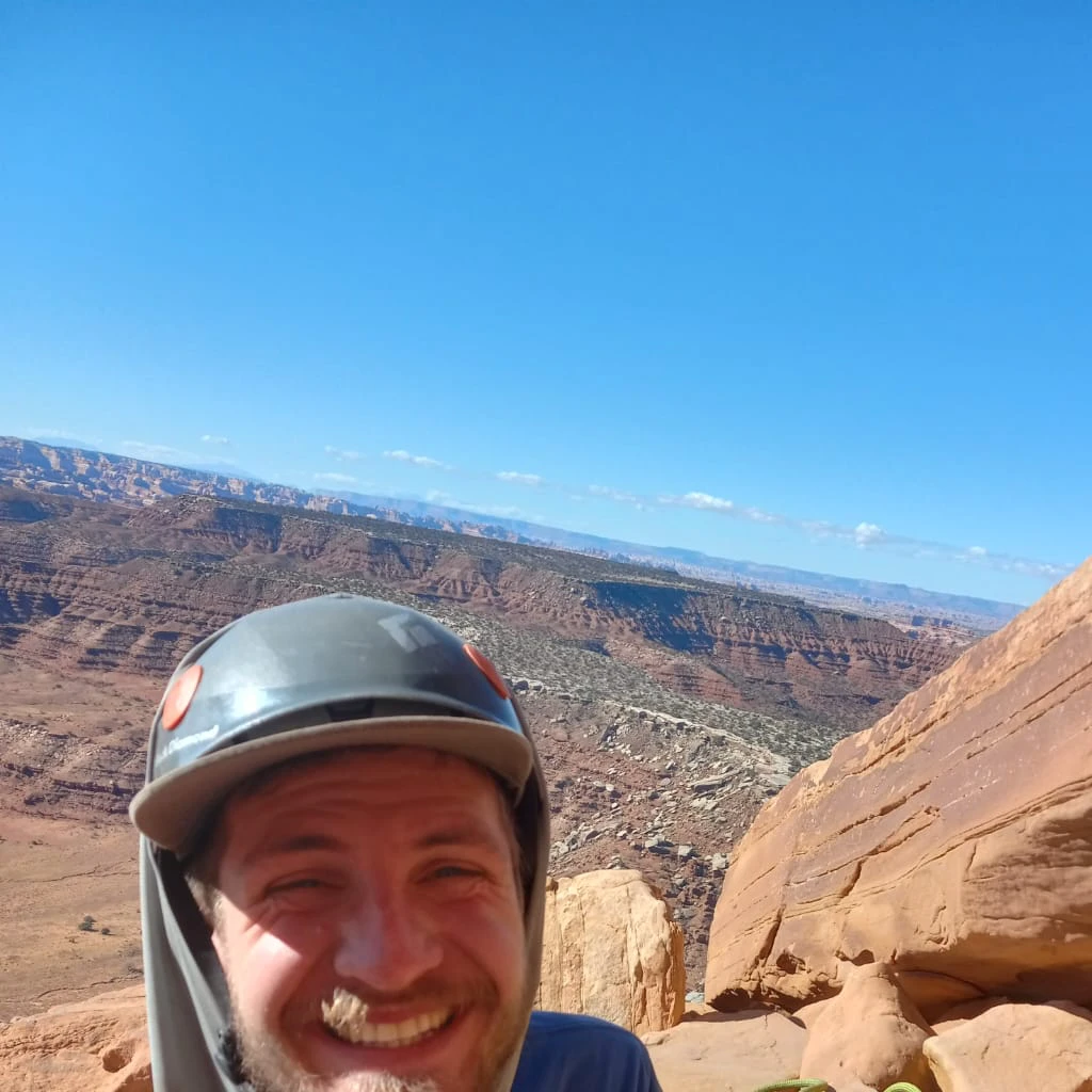smiling man in helmet and blue shirt sitting on rock with green plant, moab, at the top of a red rock canyon, rob gonsavles, view from the top, belaying, sitting atop a dusty mountaintop, smiling down from above, at the top of a mountain, helmet view, ben ridgway, by Jeffrey Smith, on top of a mountaintop, it is his birthday,