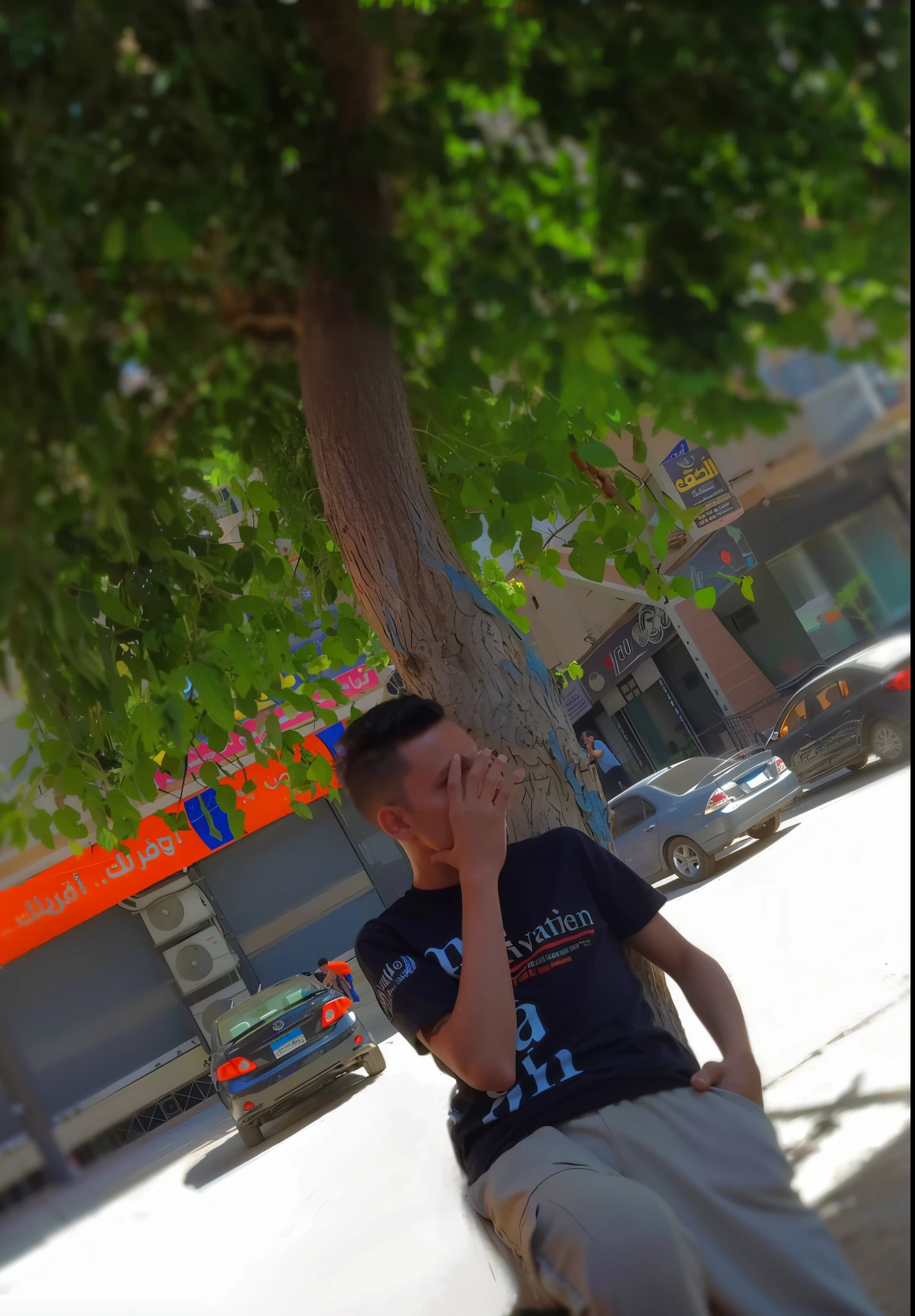 There is a man sitting on a tree trunk and talking on a cell phone, Street pic, mohamed chahin style, In sunny weather, In the street, In the street, Very, very low quality image, Next to a tree, Sitting under a tree, by Ahmed Yacoubi, On Madina Street, profile picture 1024px, amr elshamy