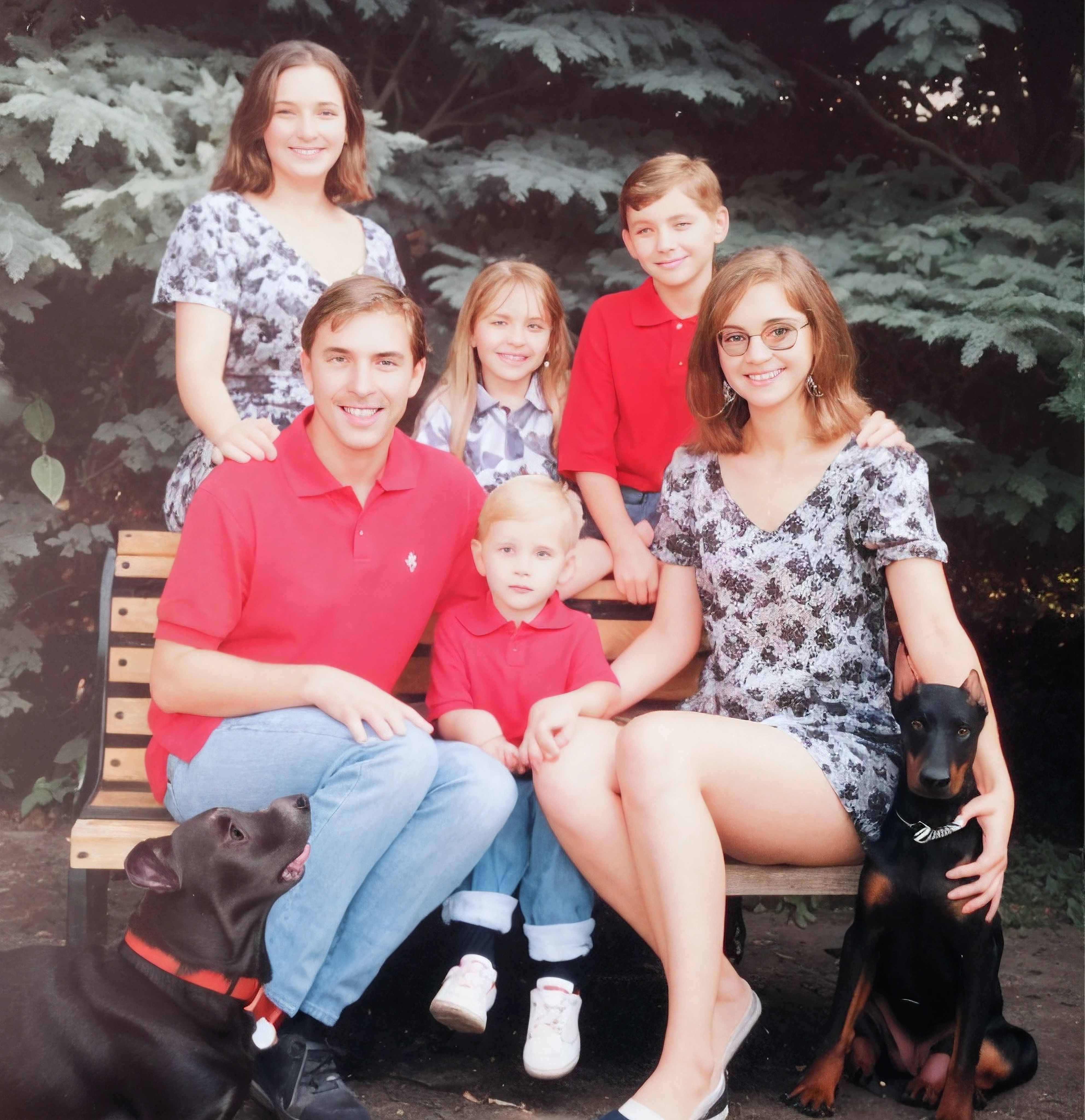 Family photo, man with light brown hair and brown eyes wearing a red polo shirt and blue jeans, meninas em vestidos cinza com desenhos de folhas pretas, meninas com cabelos castanhos, meninos com polo vermelha e jeans azul, WHITE AND RED SNEAKERS , 2 cachorros na foto, Doberman preto e marrom com coleira prateada, outro cachorro preto com nariz branco e coleira vermelha, behind there are green bushes, they are sitting on a brown planked garden bench