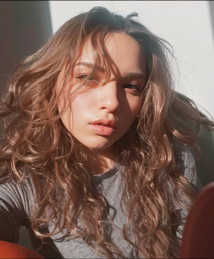 There's a woman with long hair sitting on a couch, com cabelos longos, No estilo de Petra Collins, jovem com cabelo longo, androgynous person, tirada na hora dourada, foto de perfil, com longos cabelos cacheados, androgynous face, beautiful androgynous girl, portrait androgynous girl, long messy curly hair, hard lighting!, foto do perfil