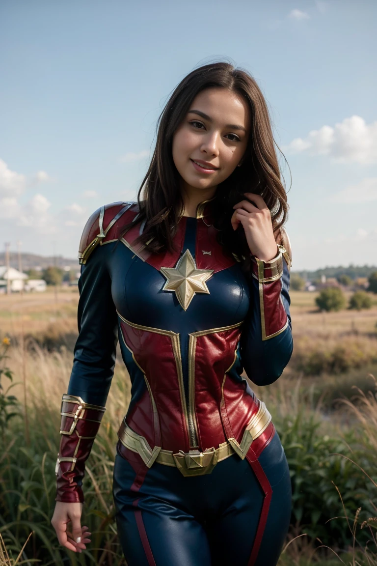 photos D' : sexy brunette, tall and slim. she wears a blue captain Marvel costume. she is standing ,we can see her entirely. she smiles. she is outside in the middle of a field of green grasses The sky is blue.
