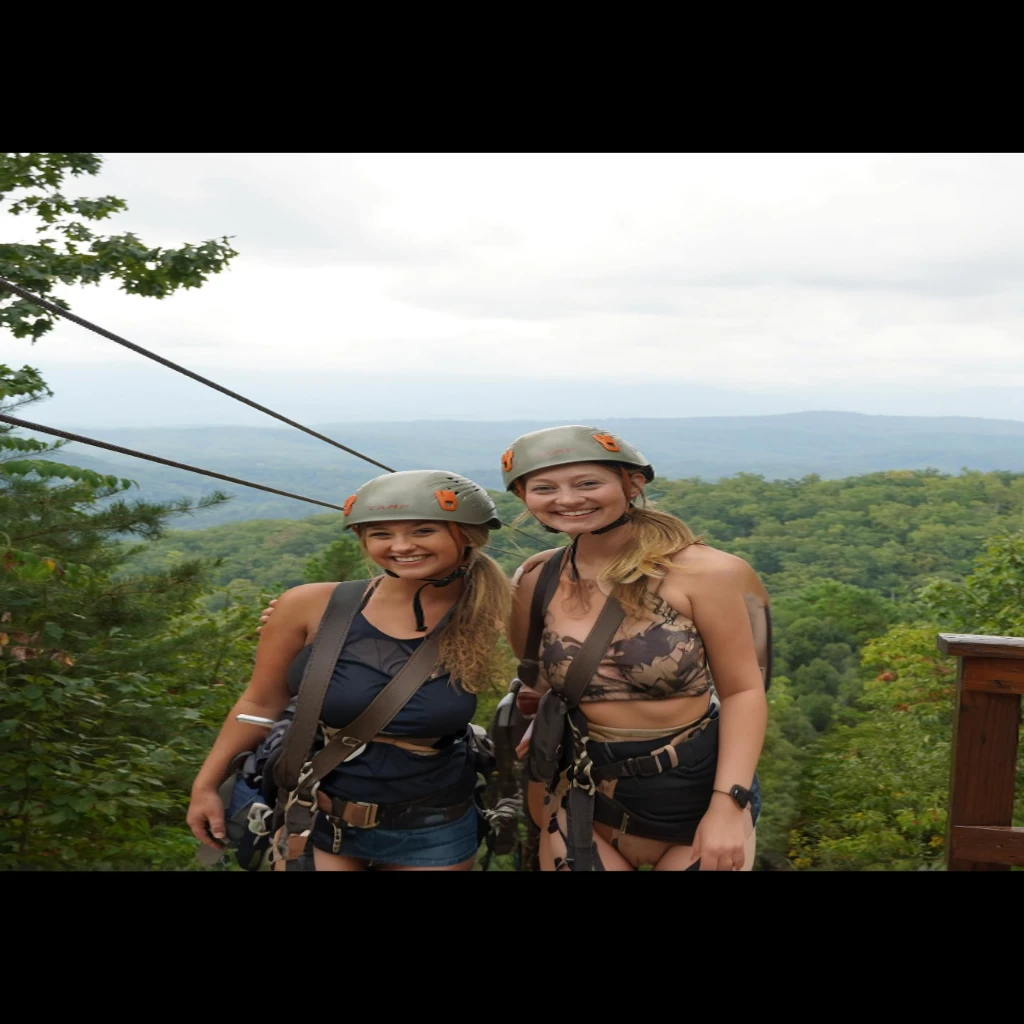 two women standing on a zipline with helmets on, over the tree tops, all in the amazing outdoors view, with mountains as background, beautiful views, with mountains in background, with mountains in the background, swinging on a vine over a chasm, mountains in the background, great view, towering high up over your view, having a great time, nude, naked,