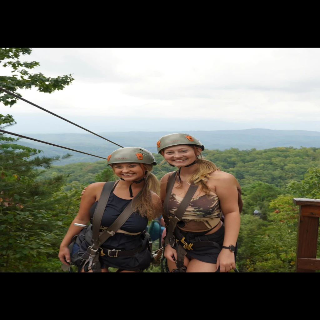 two women standing on a zipline with helmets on, over the tree tops, all in the amazing outdoors view, with mountains as background, beautiful views, with mountains in background, with mountains in the background, swinging on a vine over a chasm, mountains in the background, great view, towering high up over your view, having a great time, nude, naked,