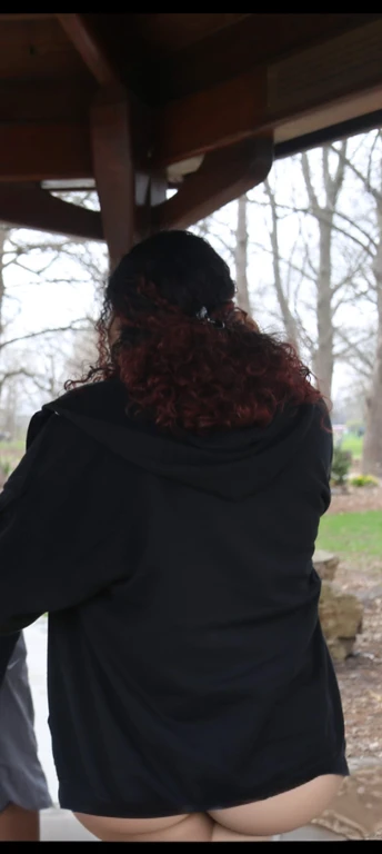 there are two people standing under a shelter talking to each other, photo still of behind view, shot from the back, view from the back, seen from the back, back of the hair, from the back, wearing a black hoodie, with curly red hair, facing away from camera, long dark curly hair, over the shoulder view, in a black hoodie, beautiful girl, big ass, huge ass, bbw, from behind, thick, thick thighs, wide hips, bbw, big tits, sideboob, giant ass, back view, big butt, round butt, tight underwear, underwear halfway off, underwear taking off, grabbing ass, ass spilling over underwear, too thick for clothes, looking left, leftwards, latina, from behind