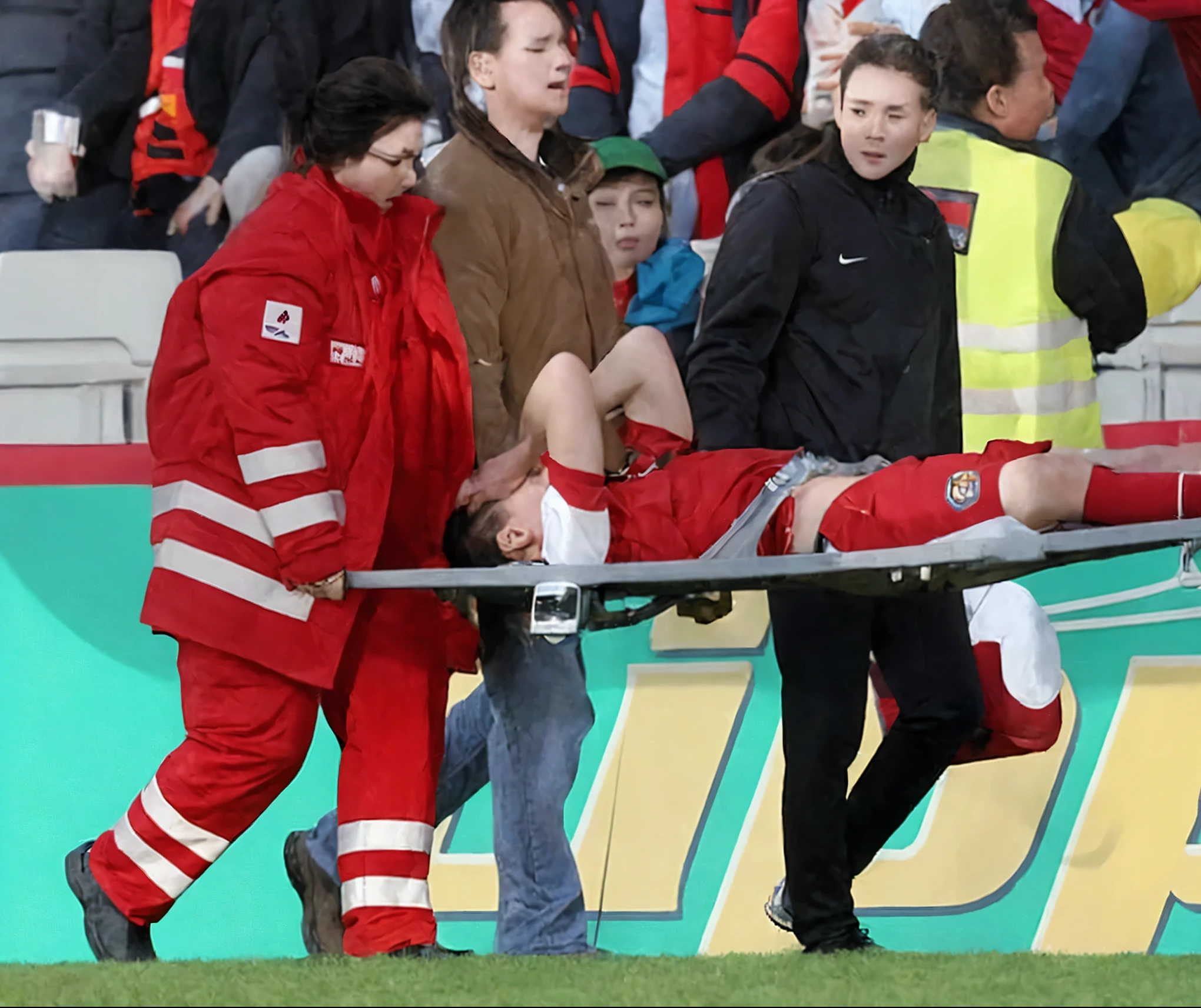 A scene at a soccer stadium in Mongolia, There is an attractive and heavily made-up Mongolian female police officer in a long black leather coat, A Mongolian female police officer in a shiny black leather coat on a soccer field, A Mongolian female police officer is holding a stretcher, A man is lying on the soccer stadium his back with his torso on the right in matt and roughened white and red sportswear on a stretcher, a man lies on a stretcher and has his left arm bent over his face, we only see the torso and thighs of the male athlete who is being carried with the stretcher to the left off the picture, the man lies on his back on the stretcher and covers his face with his bent left arm, the mongolian policewoman is exceptionally heavily made up and has an extremely desperate and very angry expression on her face, high-resolution photo, realistic sports photo, soccer scene, mongolia, mongolian policewoman, uniformed woman in black shiny long leather coat, injury scene, real photo, ultra realistic, high definition, sports photography