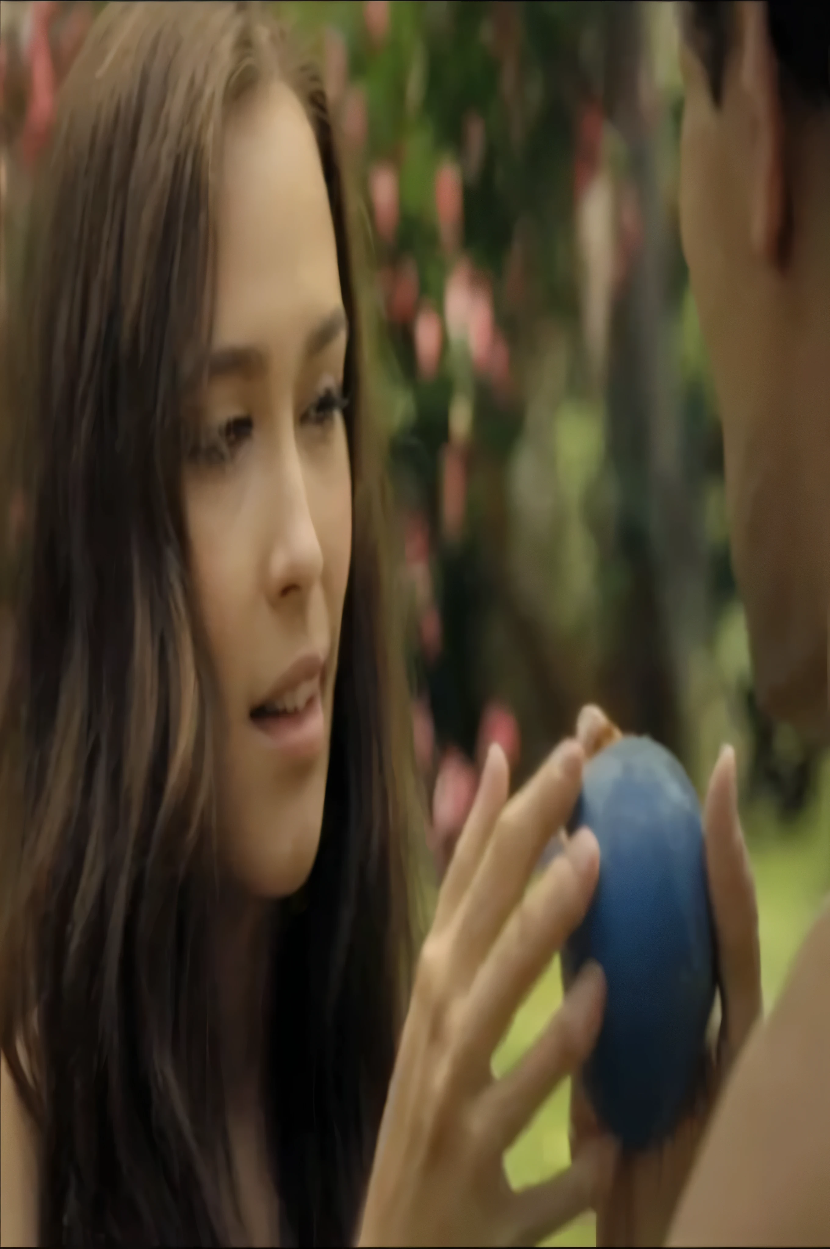 A woman and a man are in a beautiful garden, with the woman holding a golden apple. The man is looking at her as she holds the mace. They appear to be engaged in an activity or conversation involving the apple.