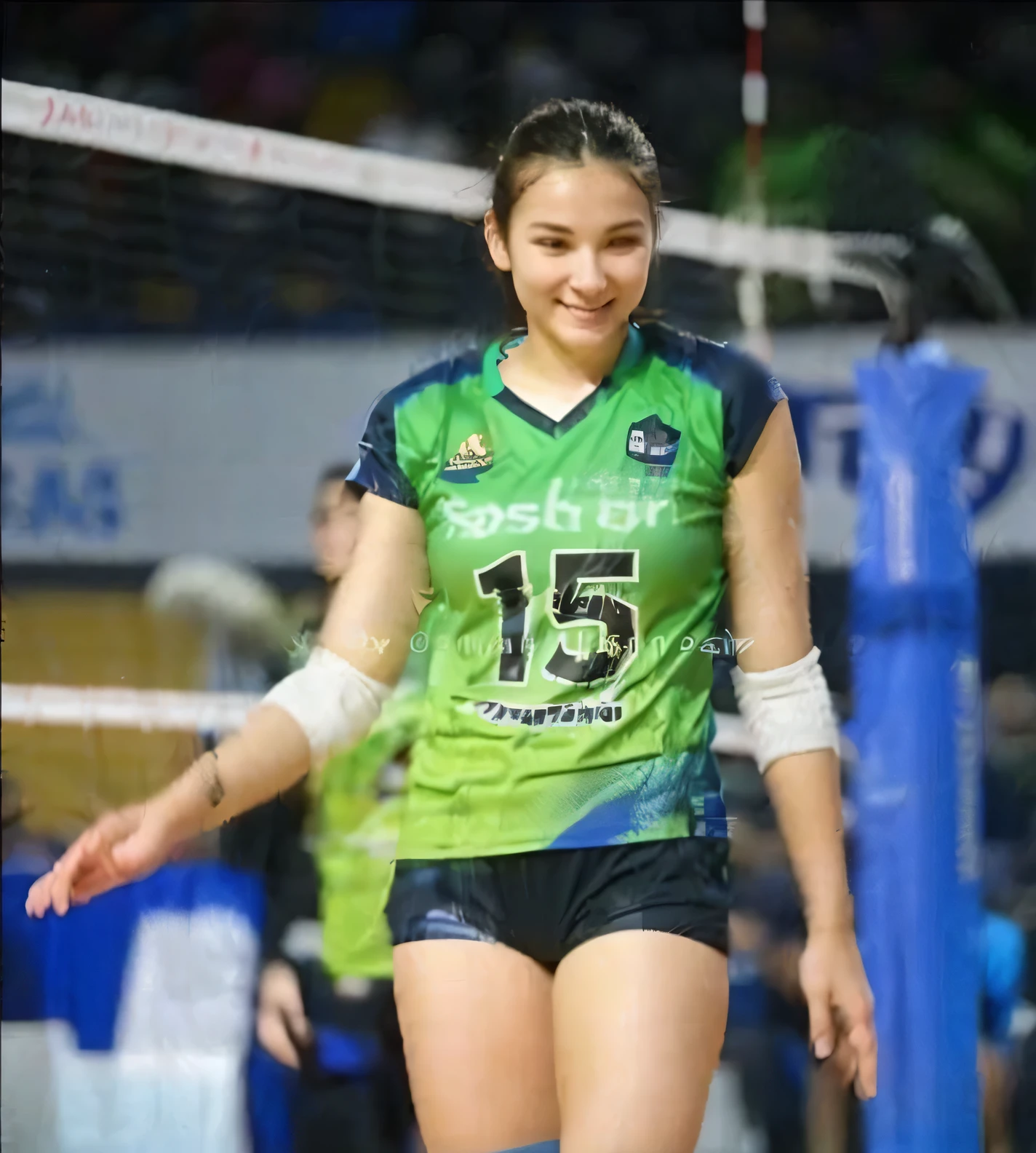 female volleyball player on the court, ideal body smiling softly and sweetly wearing a blue t-shirt with the number 15 and above it written 'JOGJA', blue shorts, black knee pads with a pink logo, volleyball net, other players dressed in red, the floor is marked with white lines