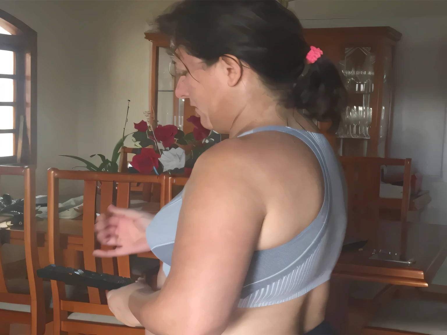 A woman stands in a dining room, facing away from the camera, wearing a gray sports bra and holding a remote control in her hand. back view, showing shoulder from behind, katanas strapped to back, 4k shot, partially cupped hands, lara croft muscle sweat, practicing her swords, flexing big muscles. She is looking at a device she holds in her other hand. The room has wooden cabinets and furniture, and on the counter there is a vase with red flowers.