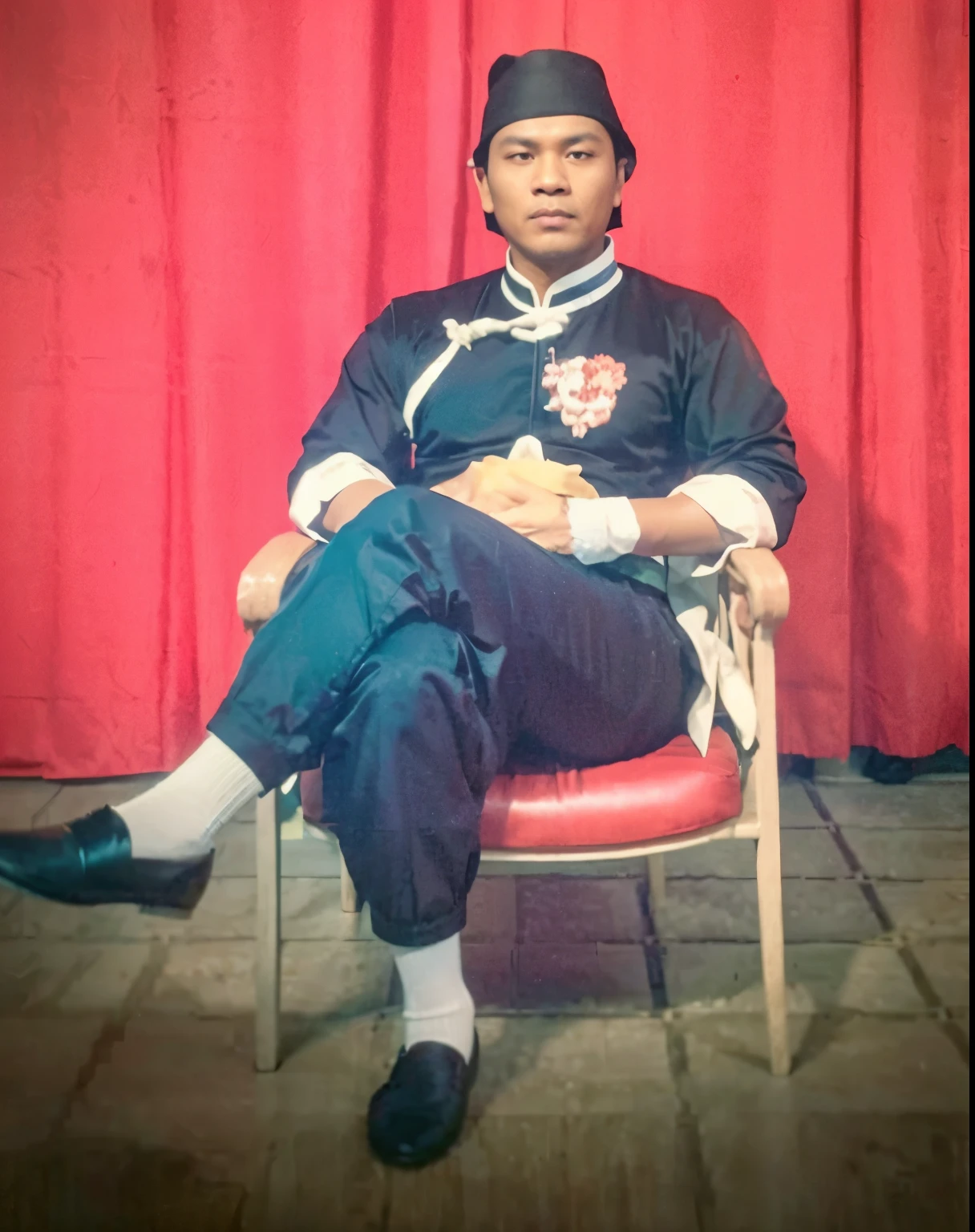 Handsome 40 year old Indonesian young man wearing a black skullcap. black cheongsam suit. long-sleeved shirt, folded sleeves, white trim. wearing a red satin belt, the ends of the belt are forked, tied at the right waist. Plain black trousers, creased cuffs. white socks, traditional kung fu slip-on shoes. Sit relaxed, leaning on a chair, near the wall, left leg crossed with the right leg. facing the camera. the whole body is visible. background inside the house. red curtain.ultra-hd.hd.65k