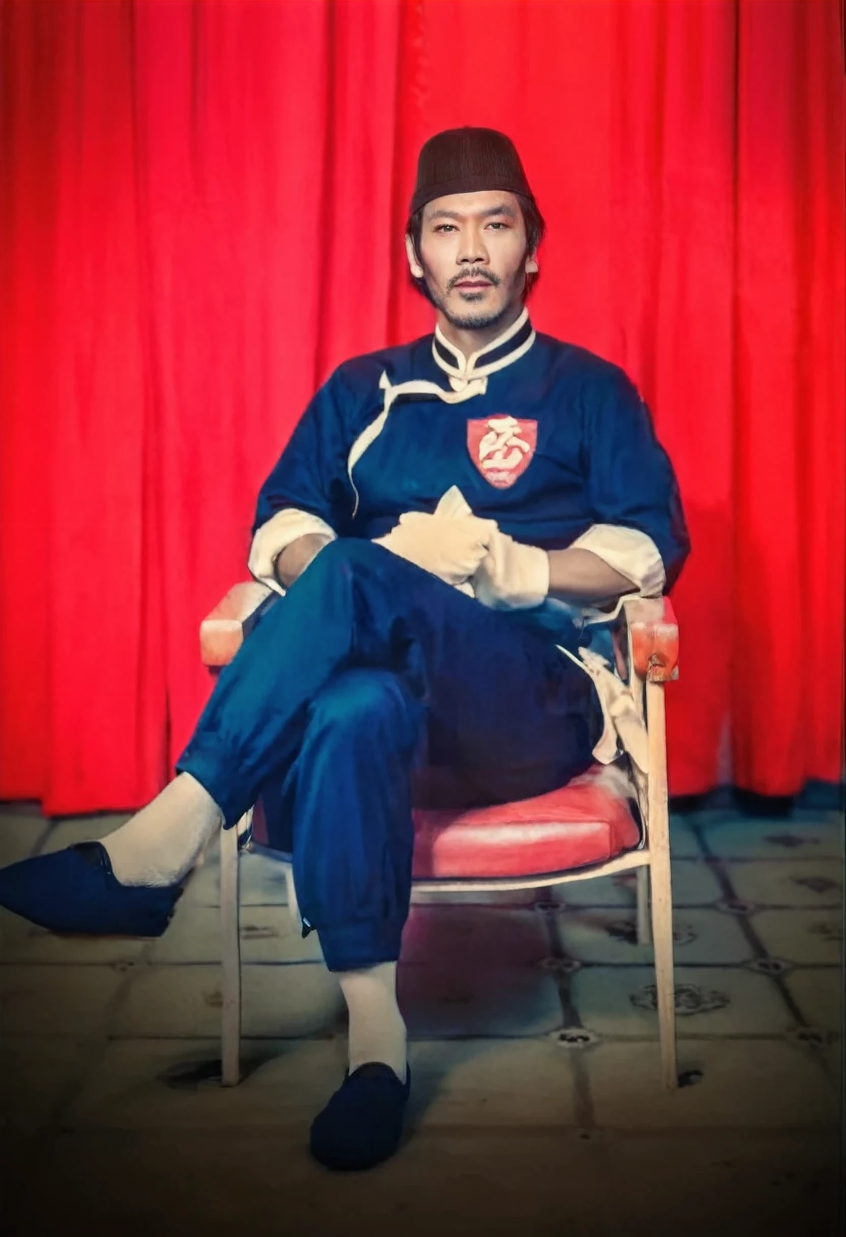 Realistic 3D caricature image of a full body Indonesian man sitting on a chair with a red curtain background. He is wearing traditional Chinese clothing, long-sleeved black cheongsam (sleeves rolled up to the elbows), shirt edges with white stripes on the edges and sleeves, shirt collar edges with stripes white. He wore a black skullcap. His hands were folded in his lap. He wore plain black joggers, white socks, and black traditional Chinese slip-on shoes (Wing Chun shoes) that matched his outfit. The man wore a calm and serious expression. The chair he sat on was red, possibly made of cloth or foam, with red armrests. He sat with his legs crossed, left leg over right. The backdrop features bright red curtains that stretch from top to floor. The floor visible below has a tile pattern, perhaps ceramic or tile, with some visible stains or dirt. Detailed HD quality cinematic 3D rendering.