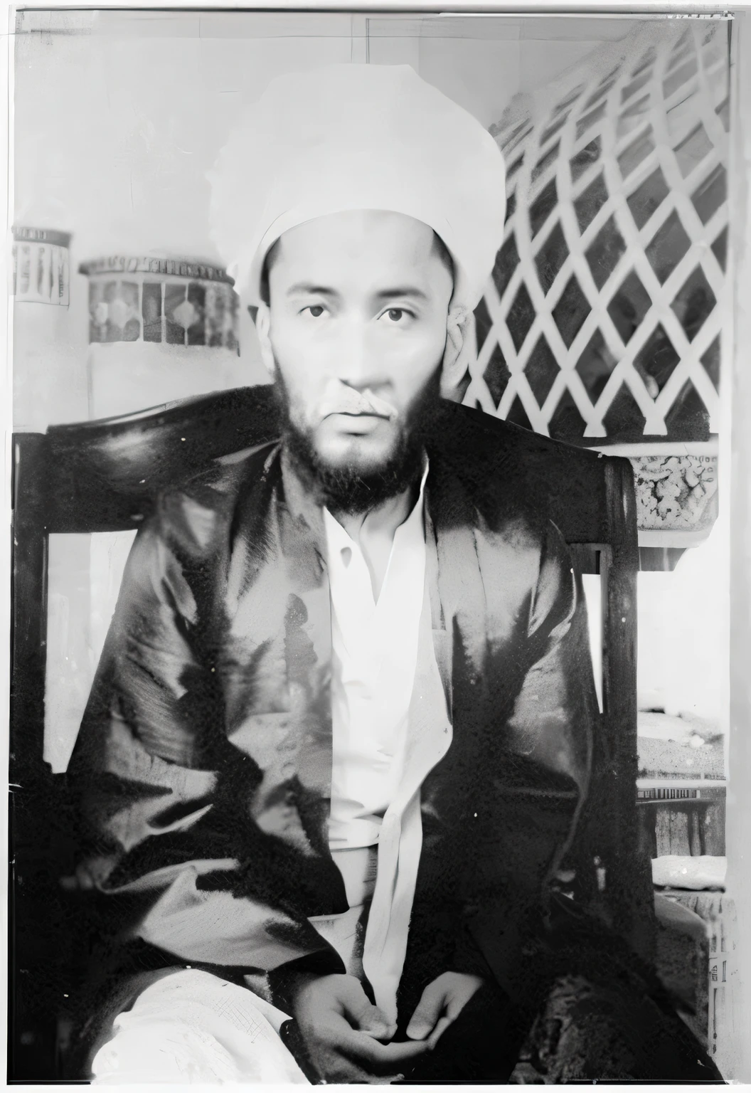 a man is sitting on a black wooden chair, looking ahead, wearing a white surjan/imama and a robe/kurta/galabiyya