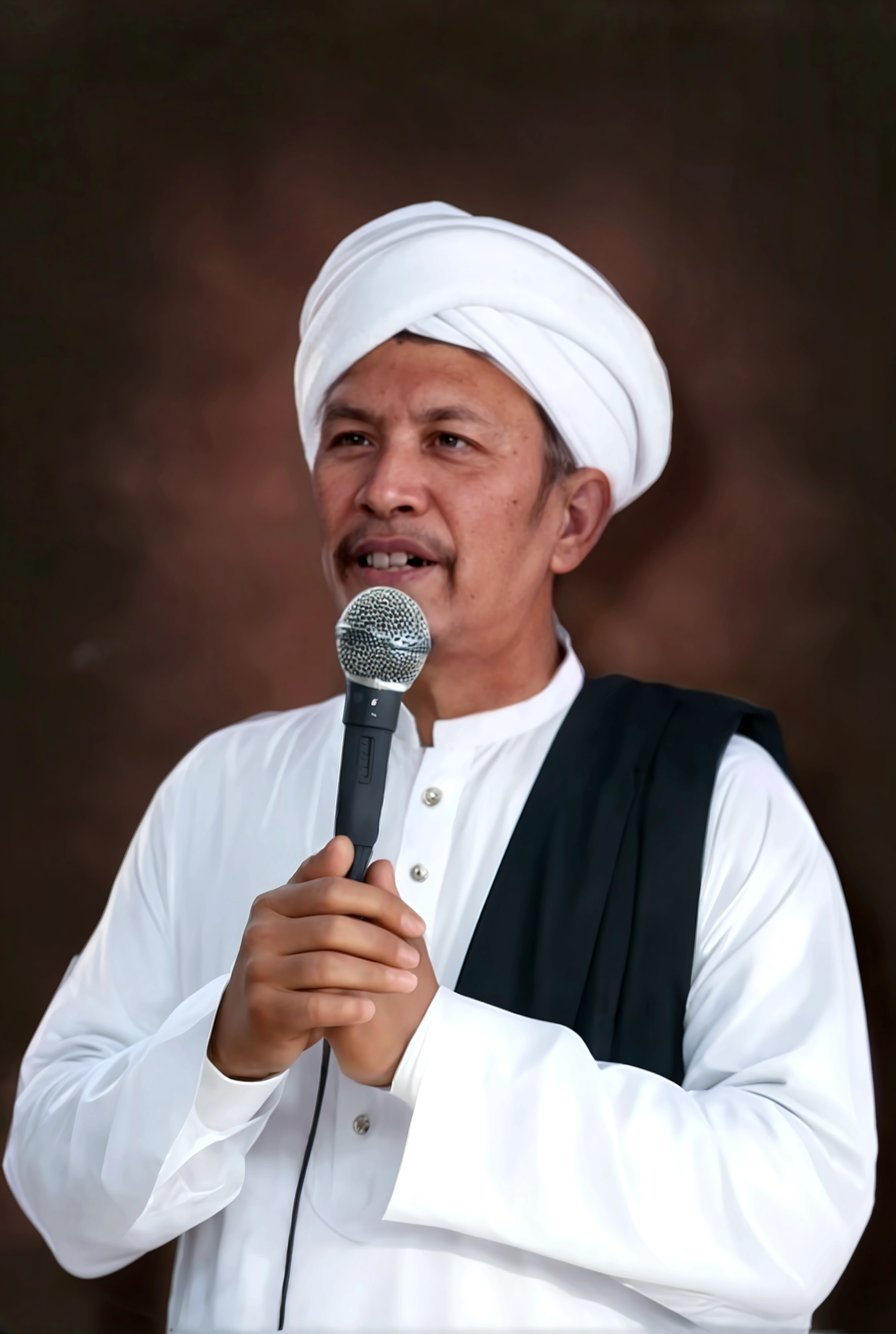 Kemeja putih, memakai sorban hijau, memegang mic, dan memakai gelang besi putih.