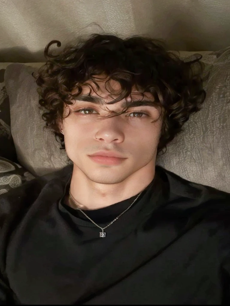 arafed man laying on a couch with a necklace on his neck, he has short curly brown hair, taken in the early 2020s, perfect handsome face, handsome face, robert sheehan, looking defiantly at the camera, inspired by John Luke, headshot profile picture, really good looking face!!, curls on top of his head, good looking face
