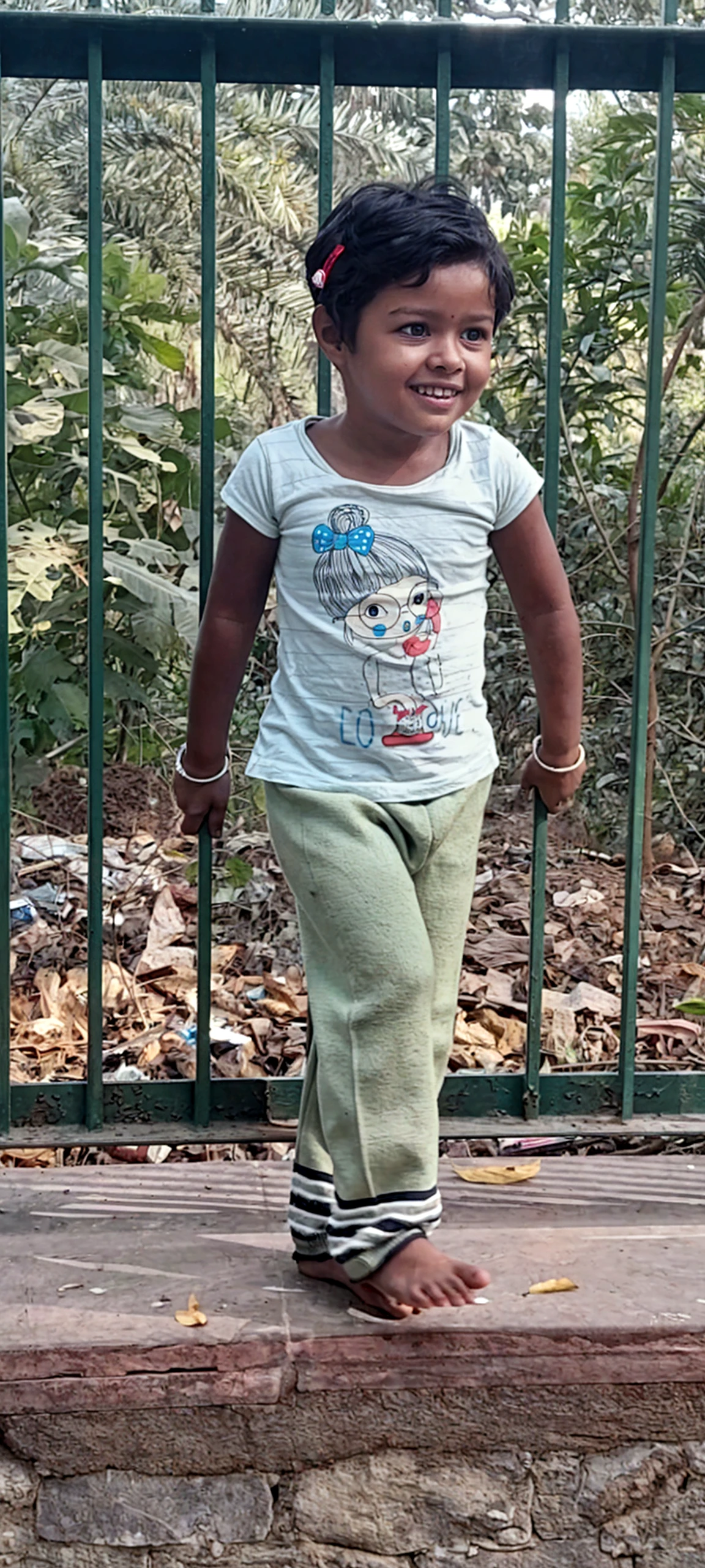 Playful and mischievous, a young woman with a jiggly fat rectangular belly and love handles, round face, olive skin, rectangular format body, small flat breasts, ponytail-hairstyle, tan skin, white shirt, denim shorts, playing with her hair, in a Latina village courtyard during the evening with a gentle sunset. In the background, a group of kids playing nearby, captured in a candid shot from a distance, with natural framing using elements in the environment.