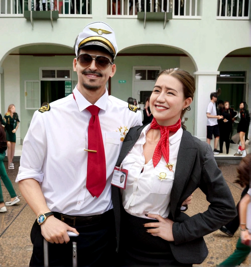 A pilot and stewardess couple, they are both smiling, o piloto esta com um relogio e uma mala em maos. The flight attendant has a red scarf around her neck and a badge hanging on her shirt.