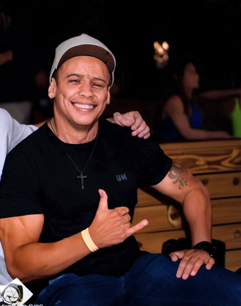 um Homen sorrindo sentado em um banco fazendo sinal de positivo usando uma camiseta preta, a light cap and blue pants, on his neck he has a chain with a cross pendant and on his left arm at the bicep he has a tattoo