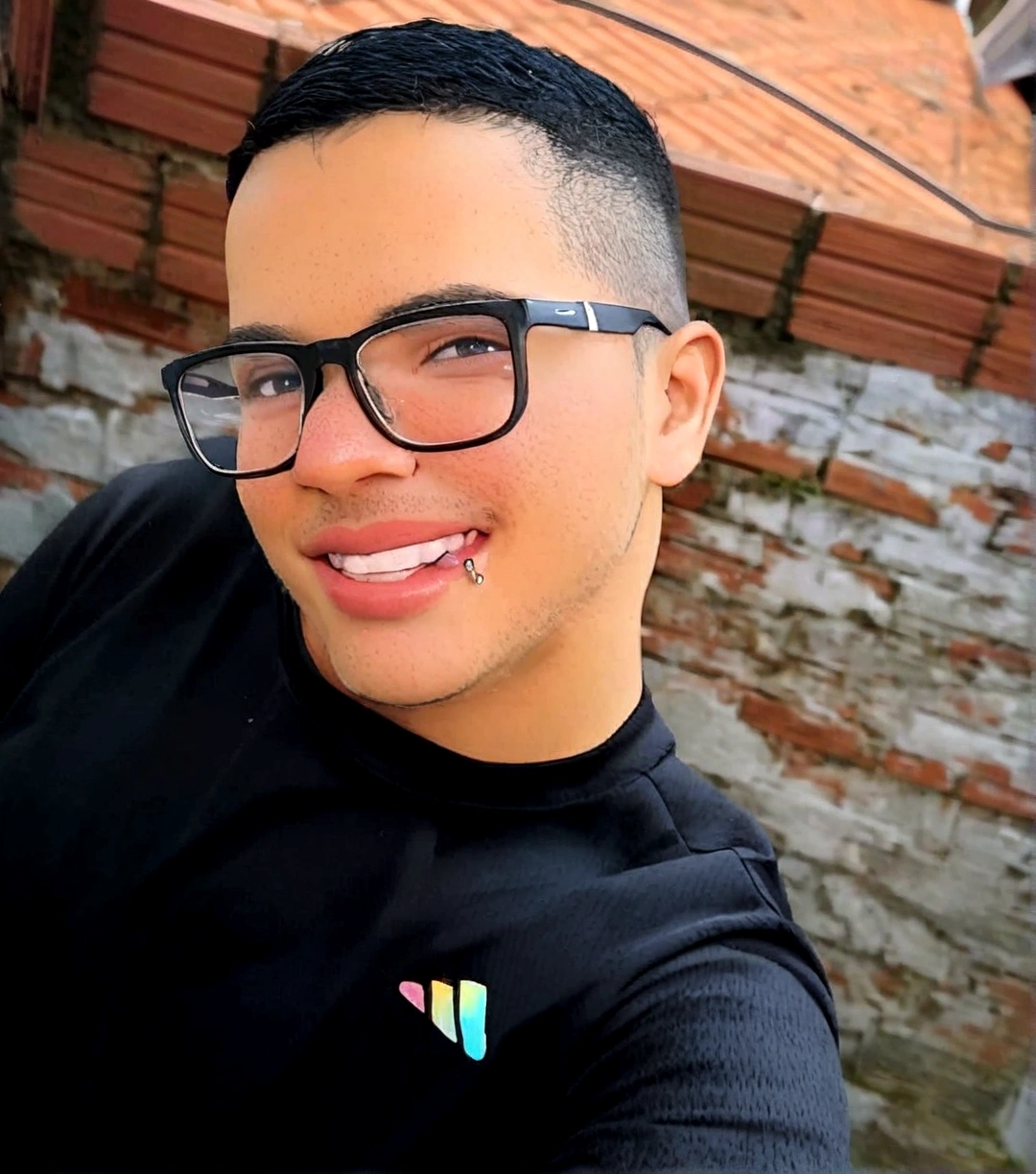 smiling man with glasses, wearing a black shirt, with a wall in the background