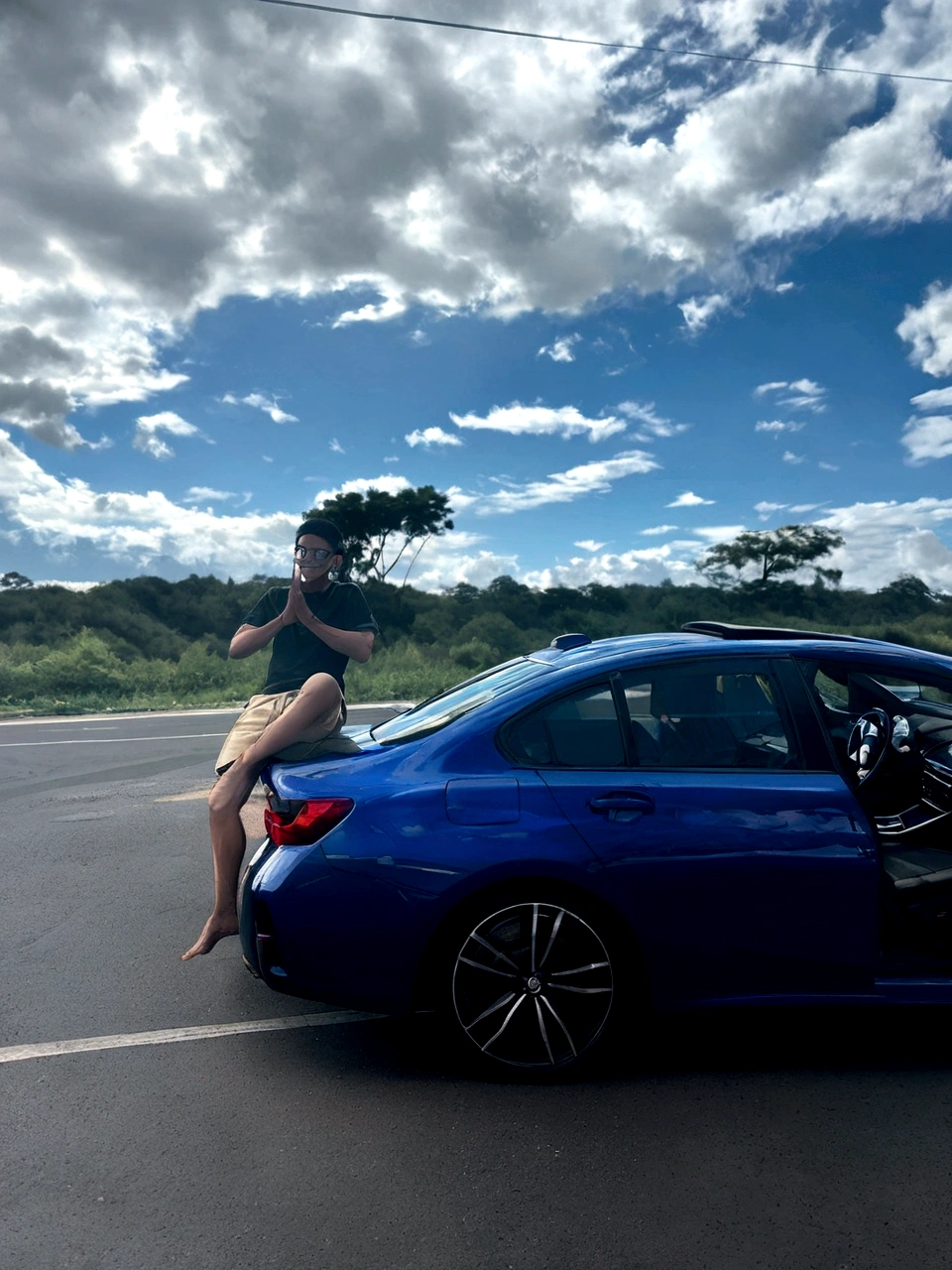 a boy smiling, on top of a car, with prescription glasses, on a road, with a very blue sky, but with clouds and a radiant sun